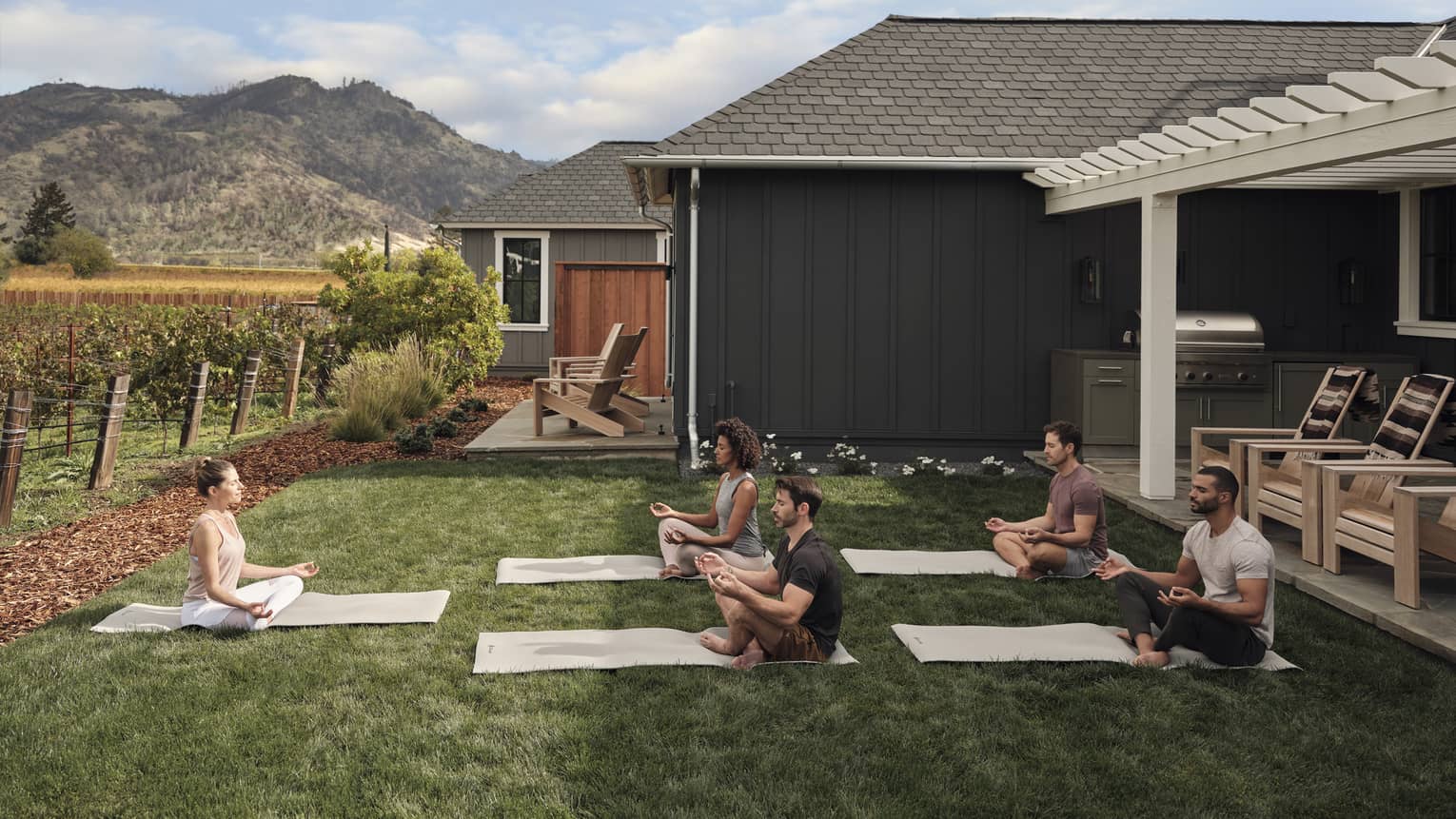 Group of people practicing yoga on mats in a grassy villa backyard near vineyards, with mountain views, wooden chairs and an outdoor grill area under a pergola