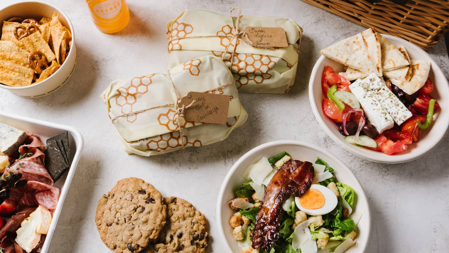 An assortment of picnic food including salad, cookies, sandwiches, fruit, and sliced meat next to a brown wood basket.