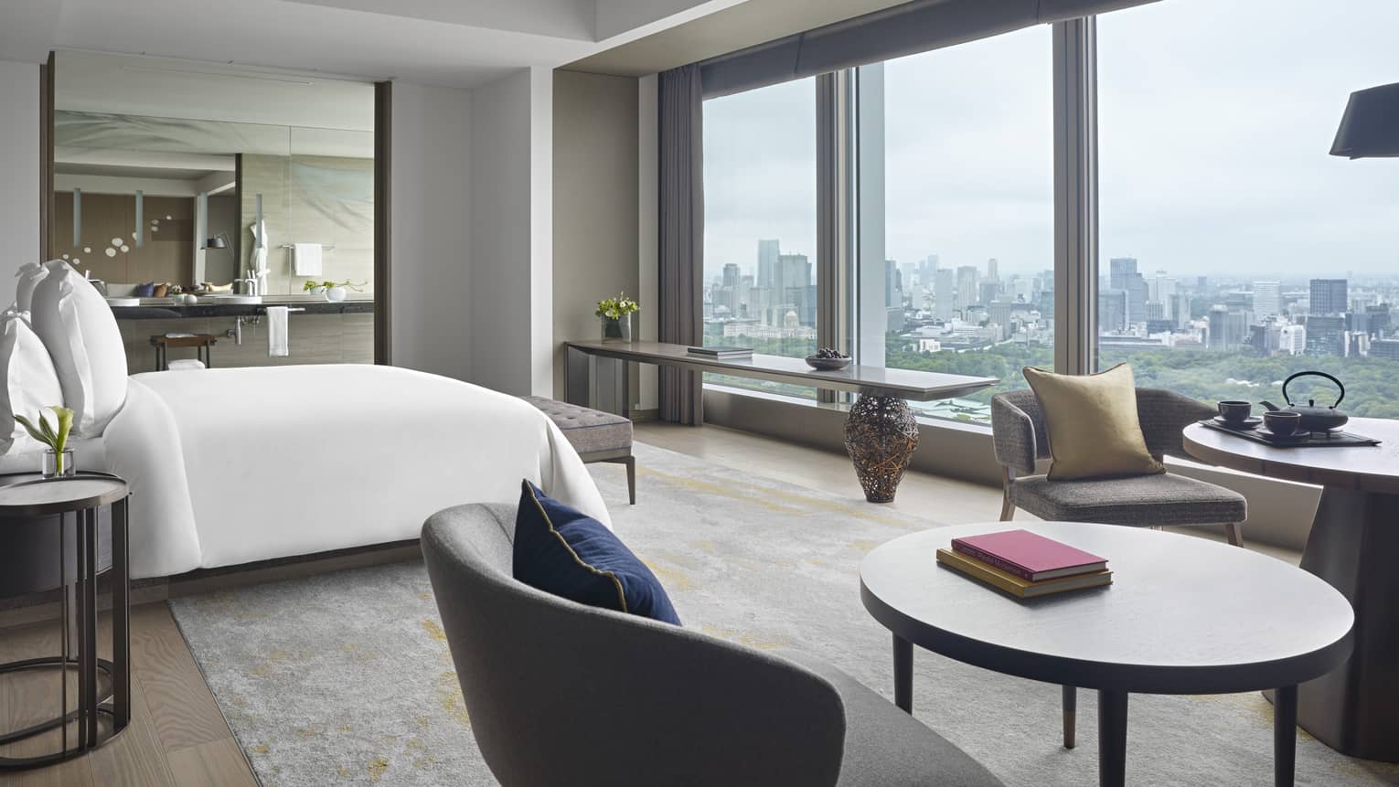 Hotel suite with a king bed, round table and chairs and a wall of windows overlooking Tokyo