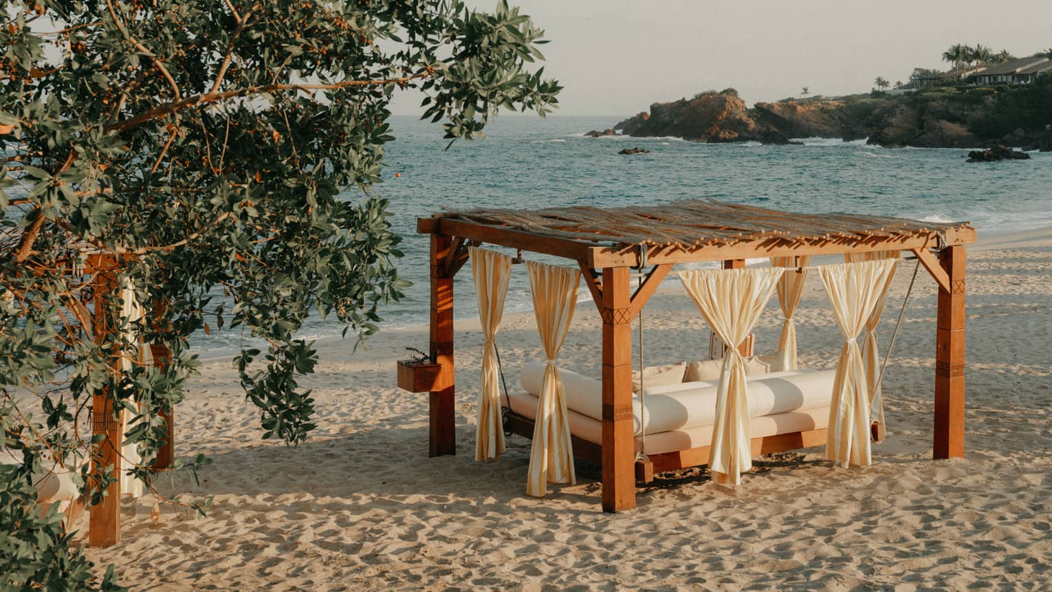 A cabana with curtains and a daybed, on a sandy beach next to stand of trees, with ocean and shoreline views in the background