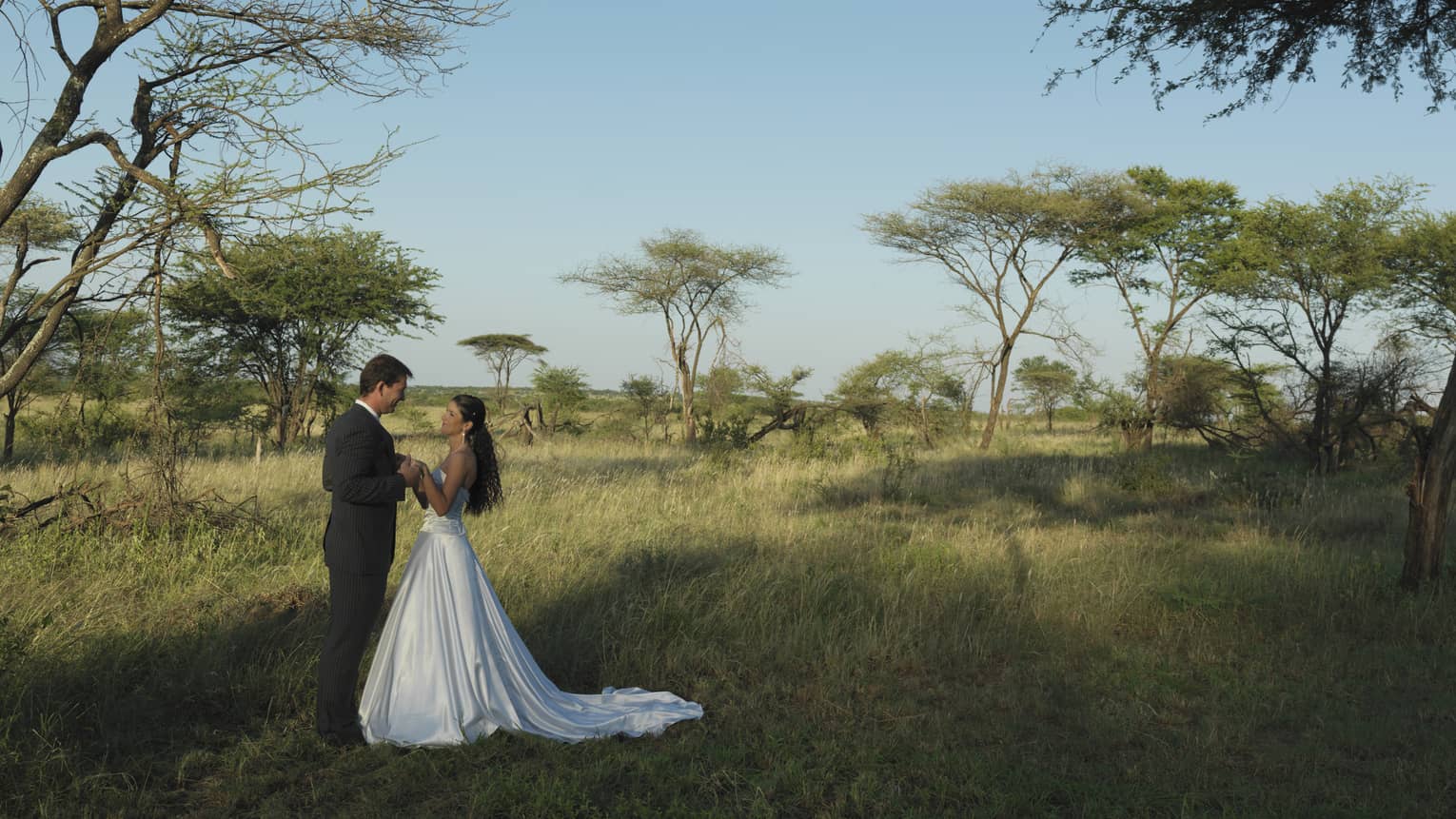 Man and woman in wedding dress hold hands standing in Serengeti surrounded by trees, blue sky