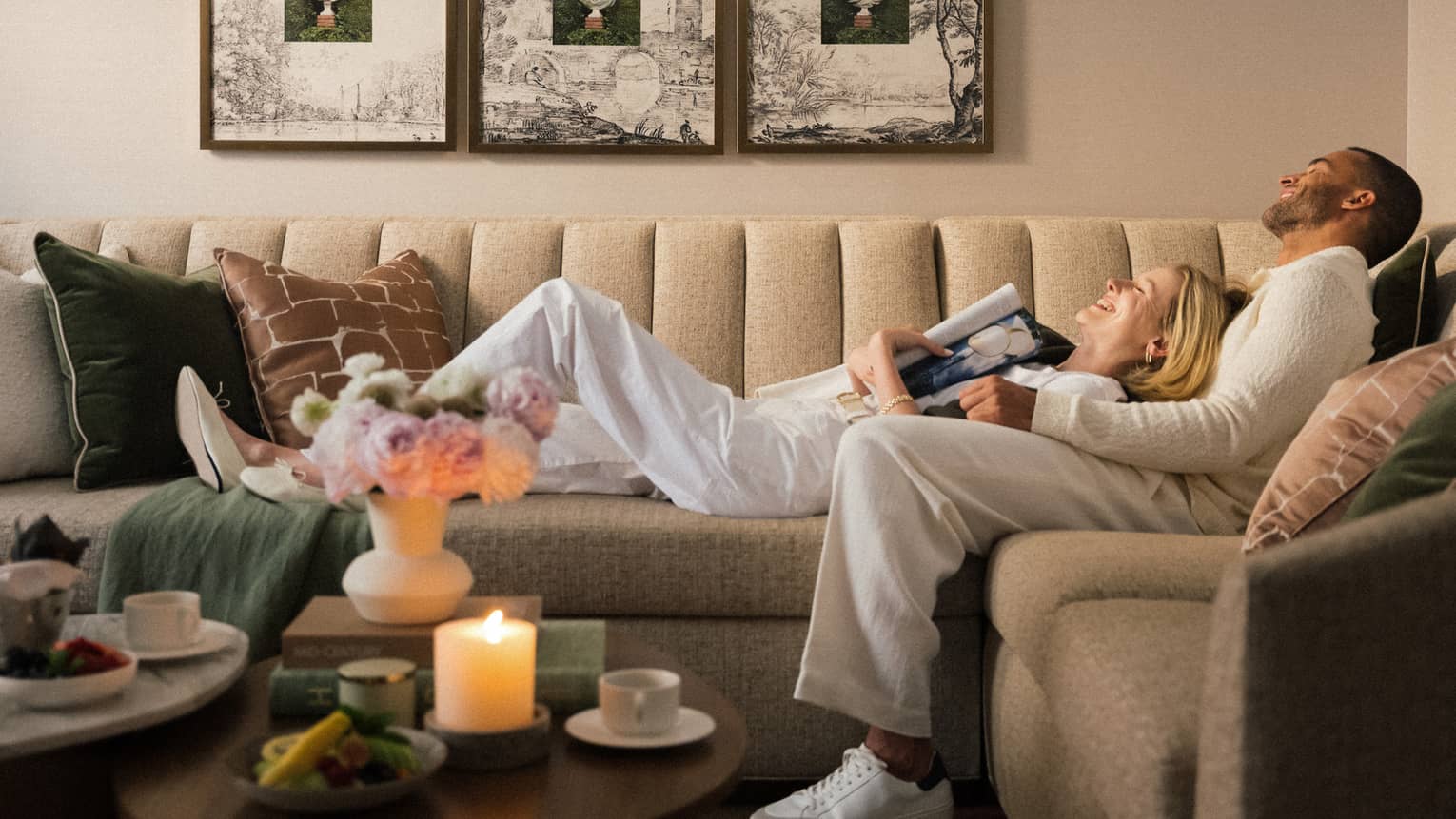 Two guests laying on the couch in a guest room with picture frames on the wall and a bouquet of flowers on the coffee table