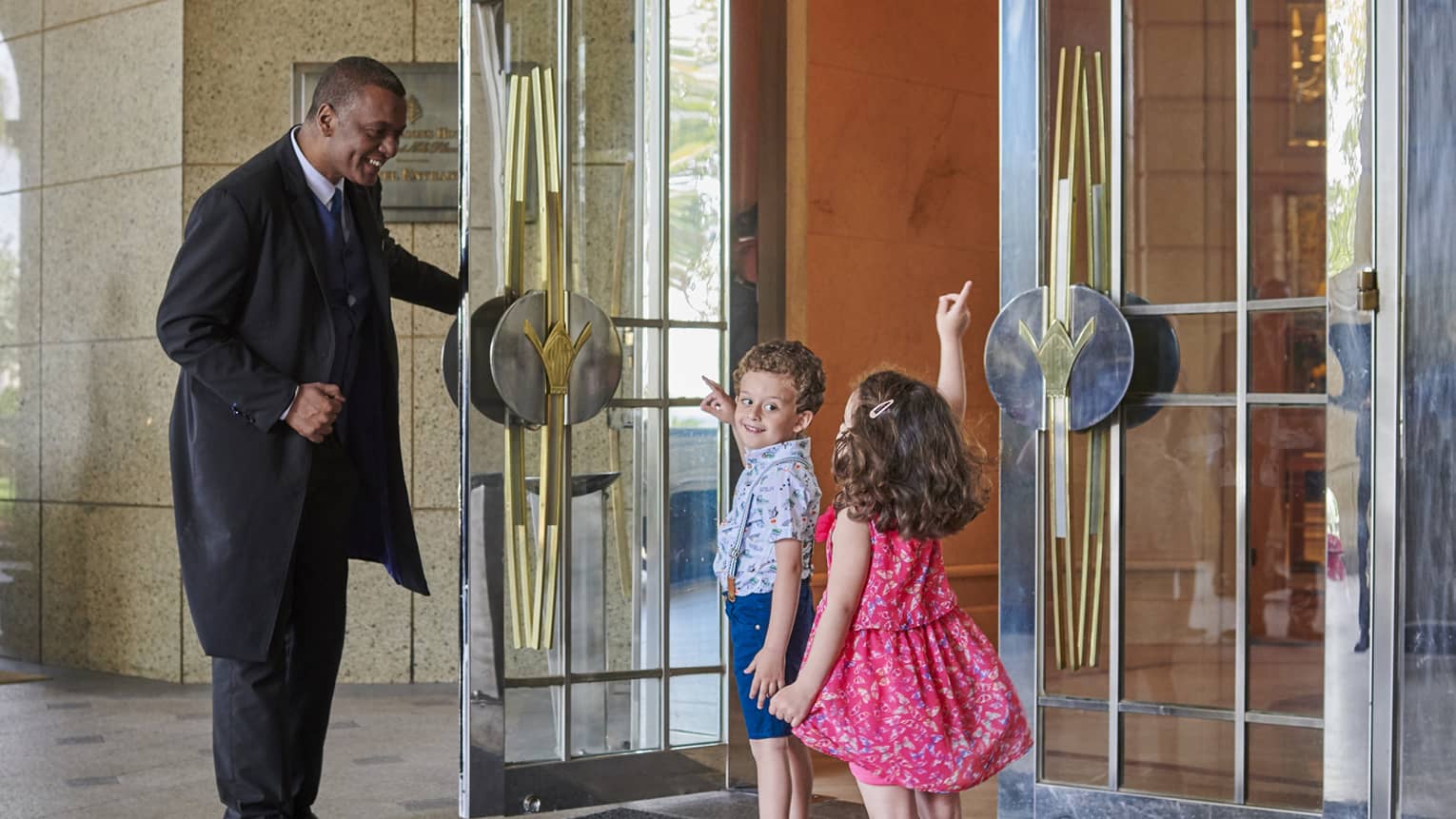 Two young children point upwards while talking to a smiling doorman wearing a morning suit who holds a door open for them.