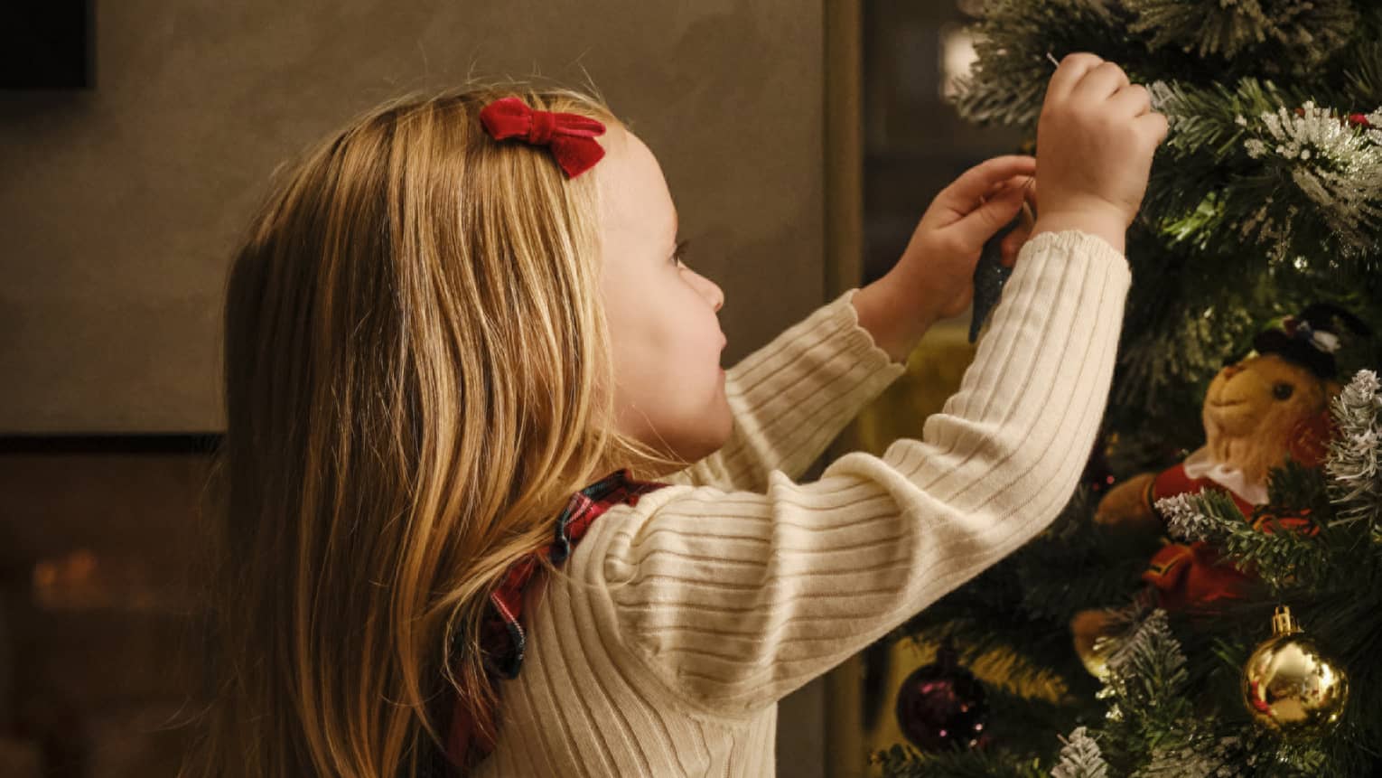 Close-up of a young child, red velvet barrette in their smooth blonde hair, arms reaching up to decorate a Christmas tree.