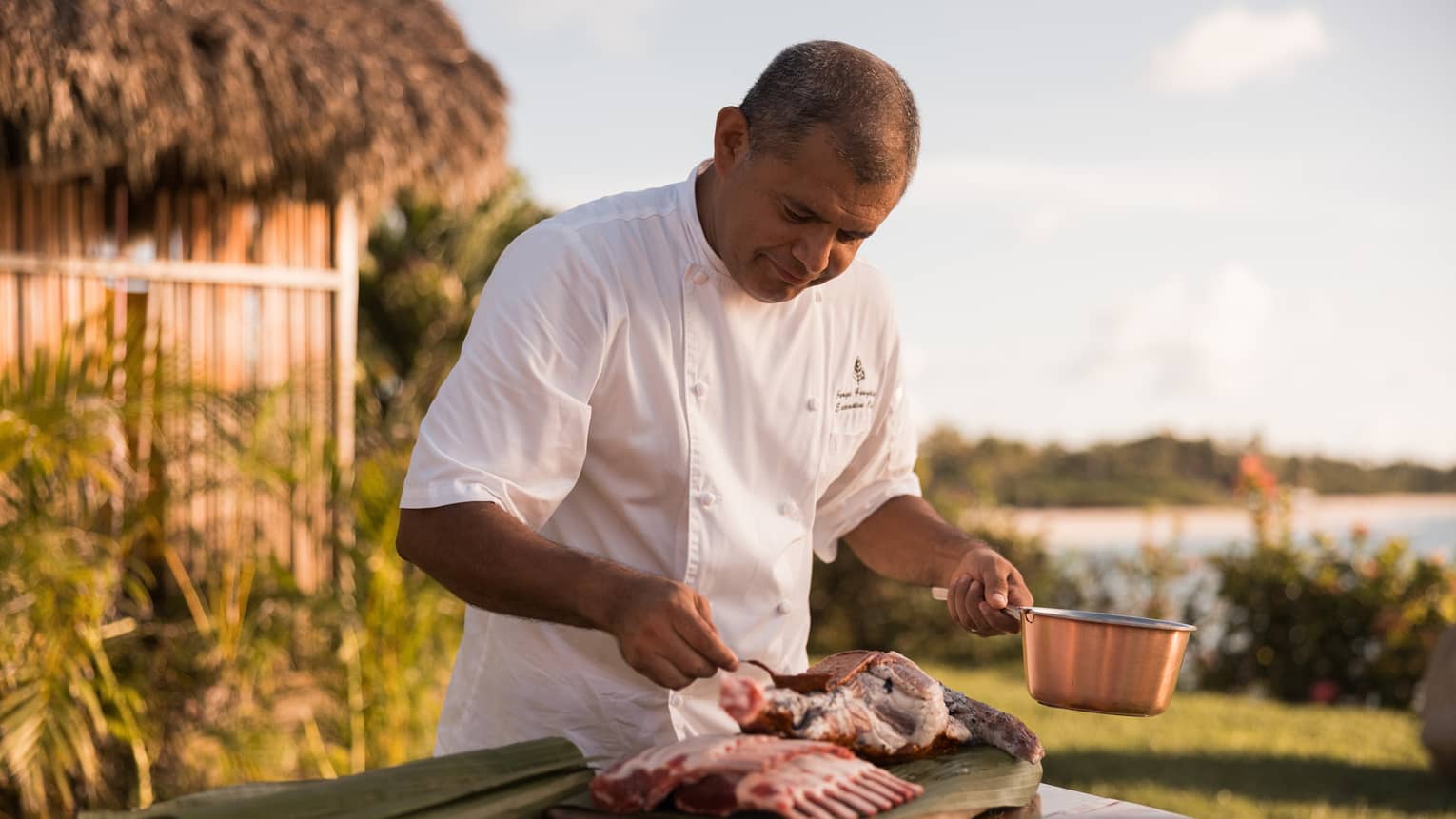 Chef in white chef’s coat, standing at an outdoor table, holding a copper pot in one hand and seasoning meat with the other