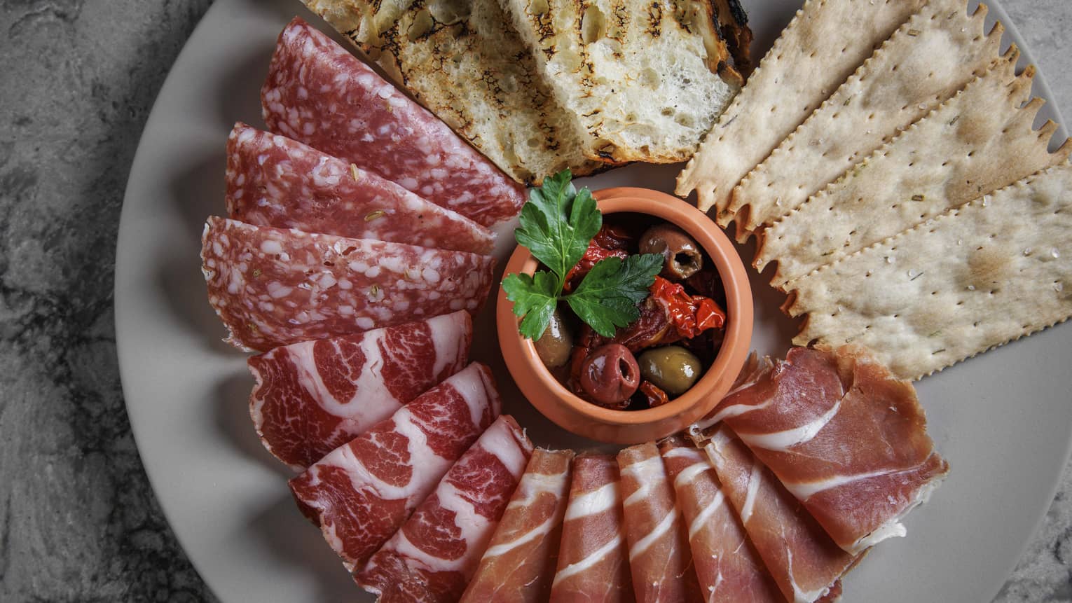 Aerial view of assorted meats, bread and crackers on grey plate with bowl of olives in the centre