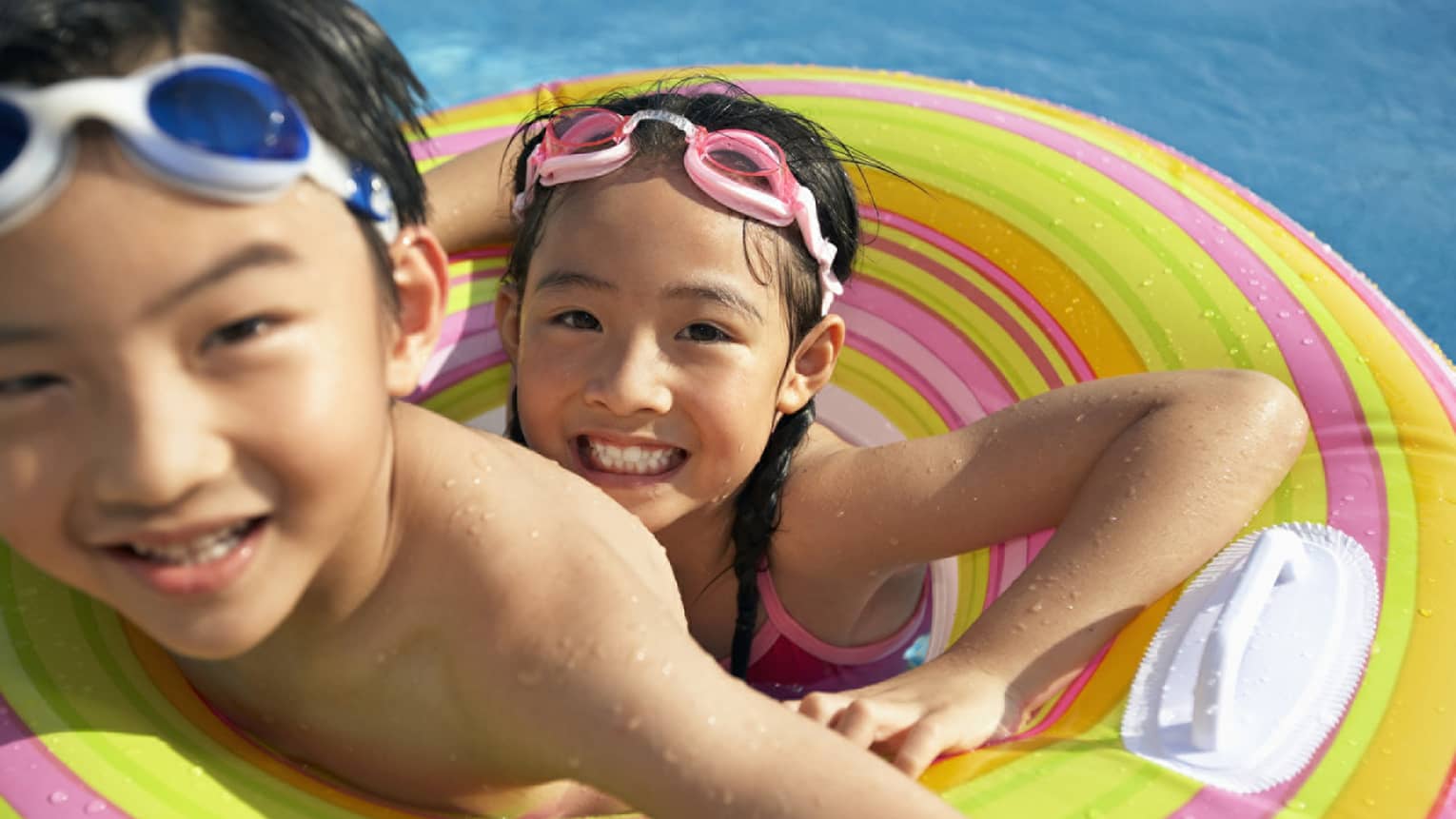 Smiling brother and sister with goggles on head share inflatable tube in swimming pool
