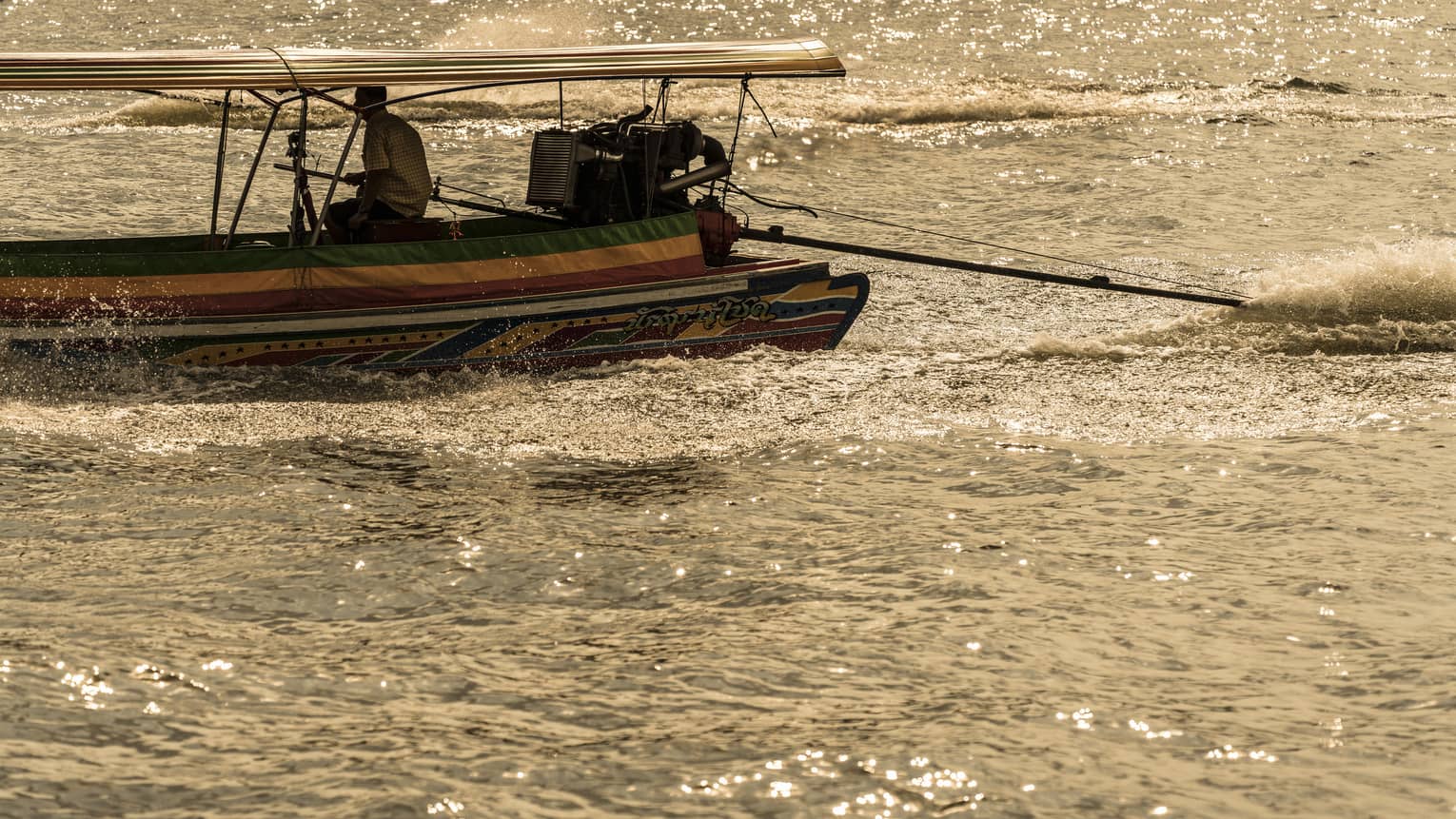 A boater sits on the edge of a colourful longtail boat, producing a frothy wake while sailing through sun-dappled water.