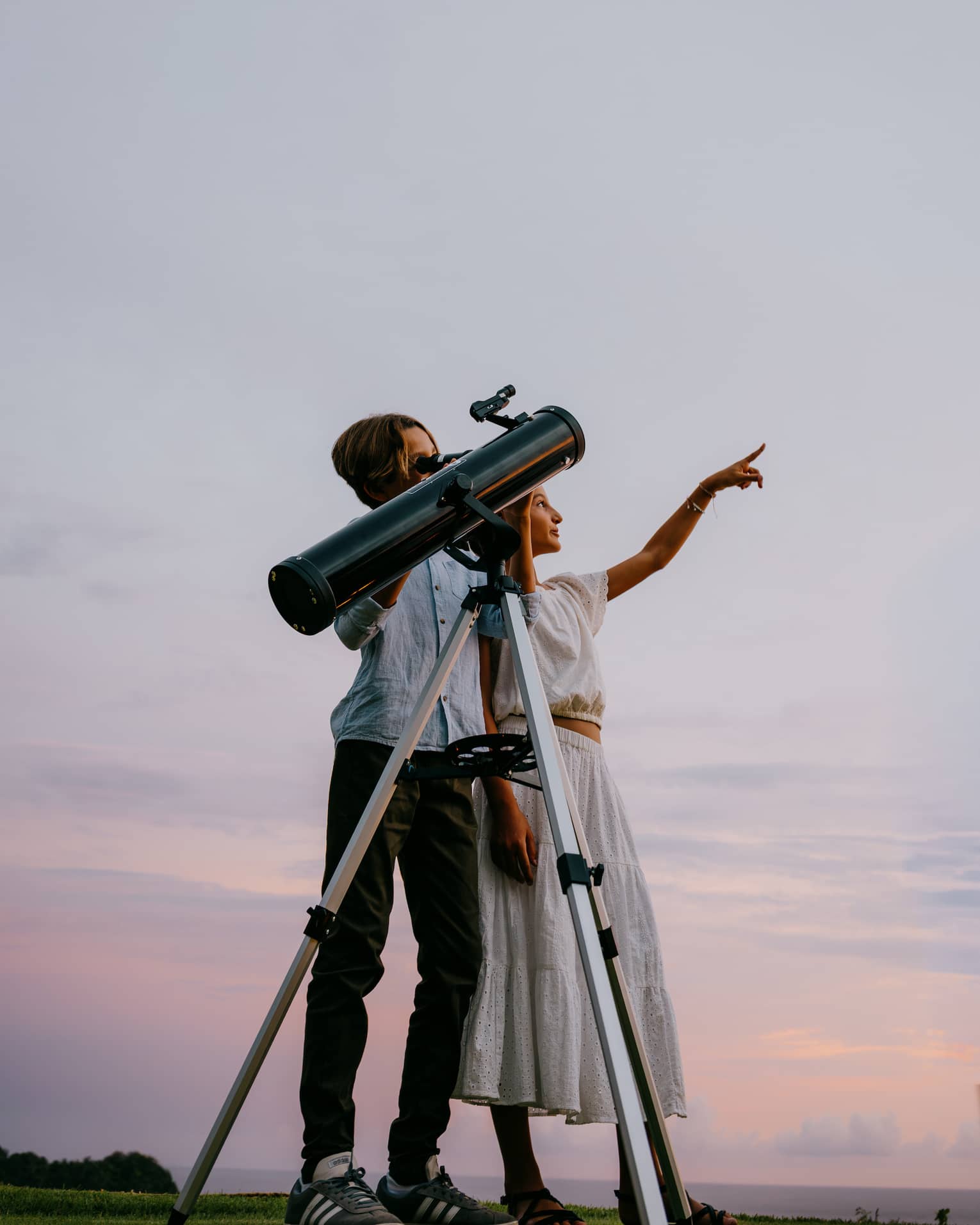 Two people stand next to a large telescope with an evening sky in the background. One person points toward the sky.