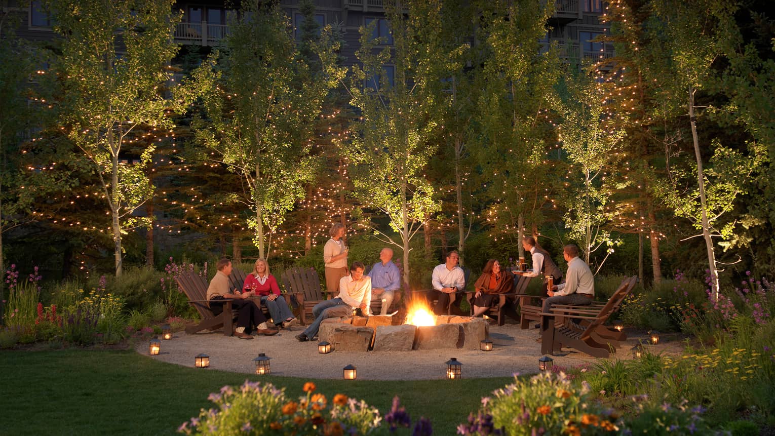 People in adirondack-style chairs with wine, cocktails around roaring bonfire, trees with lights