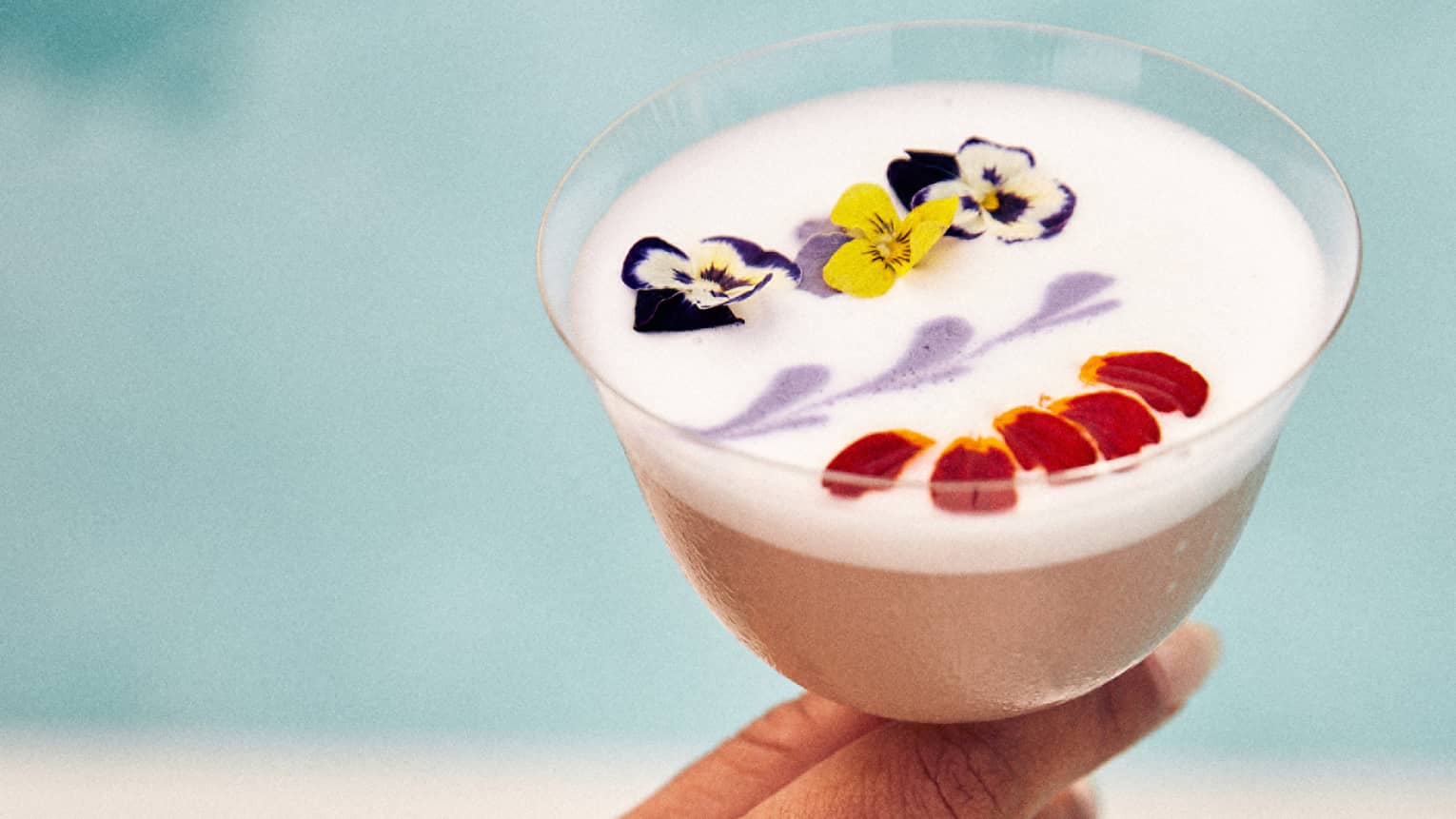 A white cocktail with flowers in it being held by a person near a pool.