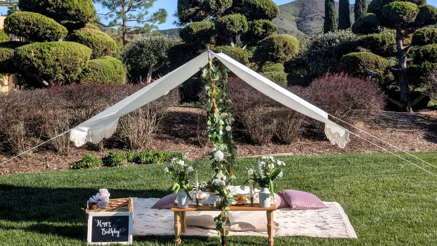 Picnic set up on a lawn with blanket, pillows, small table, flower garlands and white canopy
