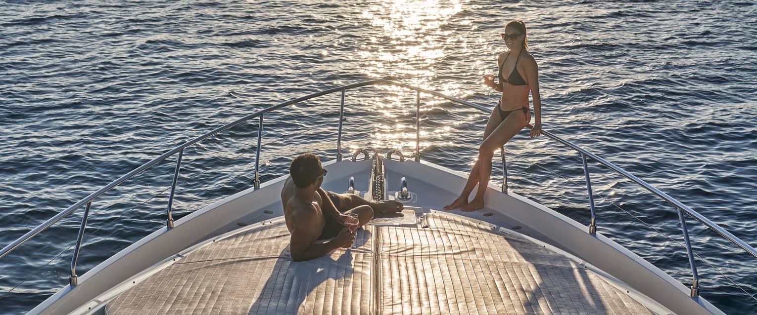 A couple in swimwear relaxing on the bow of a boat, wine glasses in hand and the setting sun glinting off the endless ocean.
