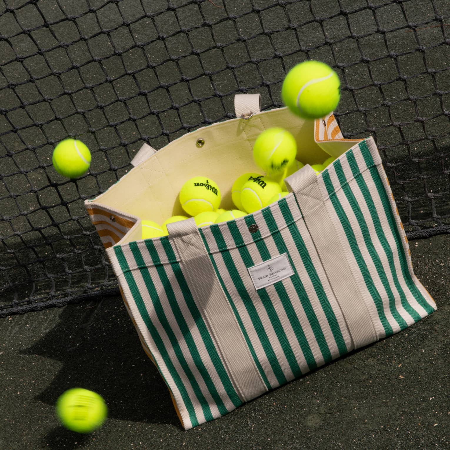 Green-and-white striped canvas bag filled with tennis balls leans against a net