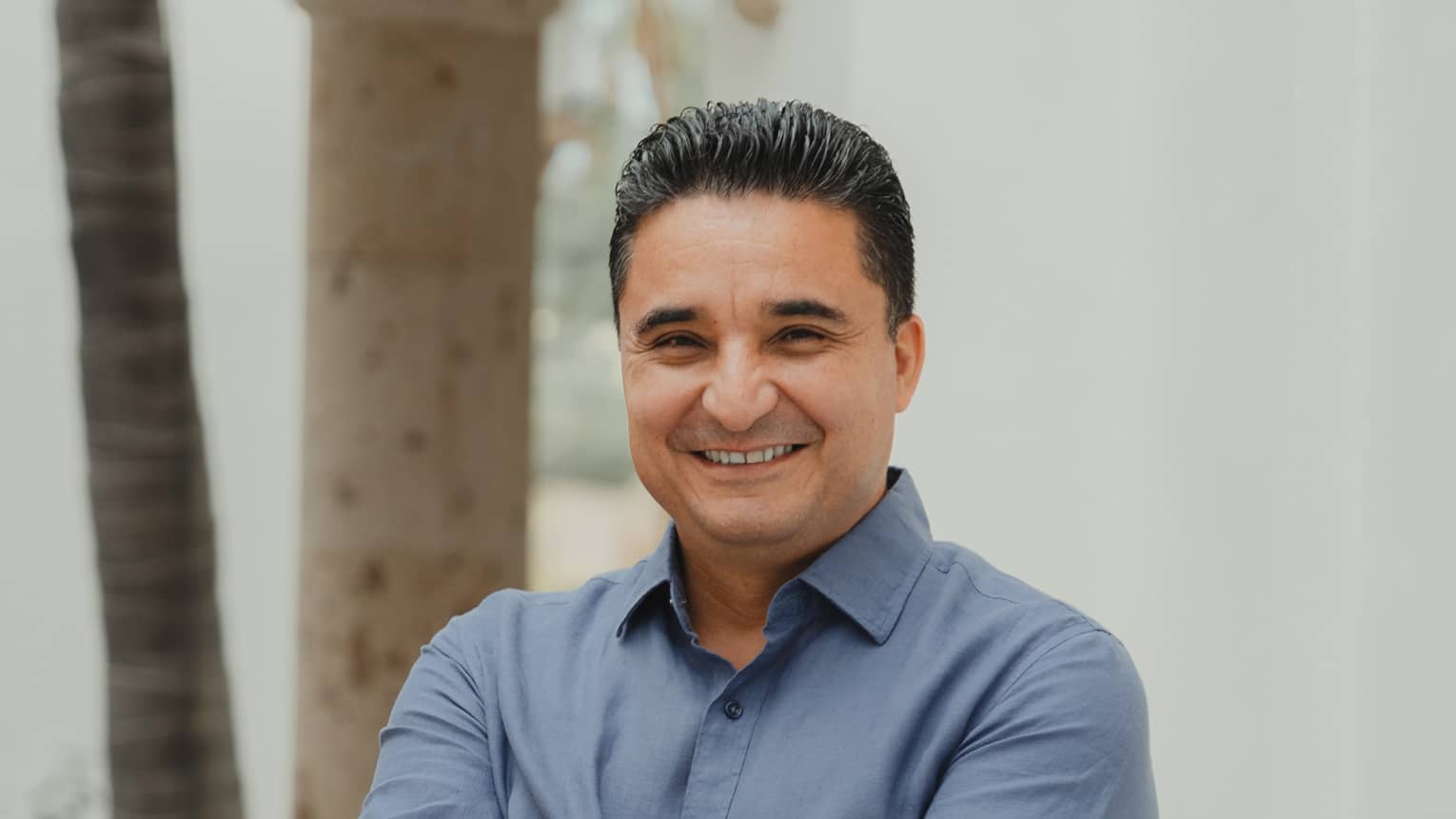 Person with short dark hair wearing a blue button-down shirt and khakis stands smiling with arms crossed in front of a white plaster wall