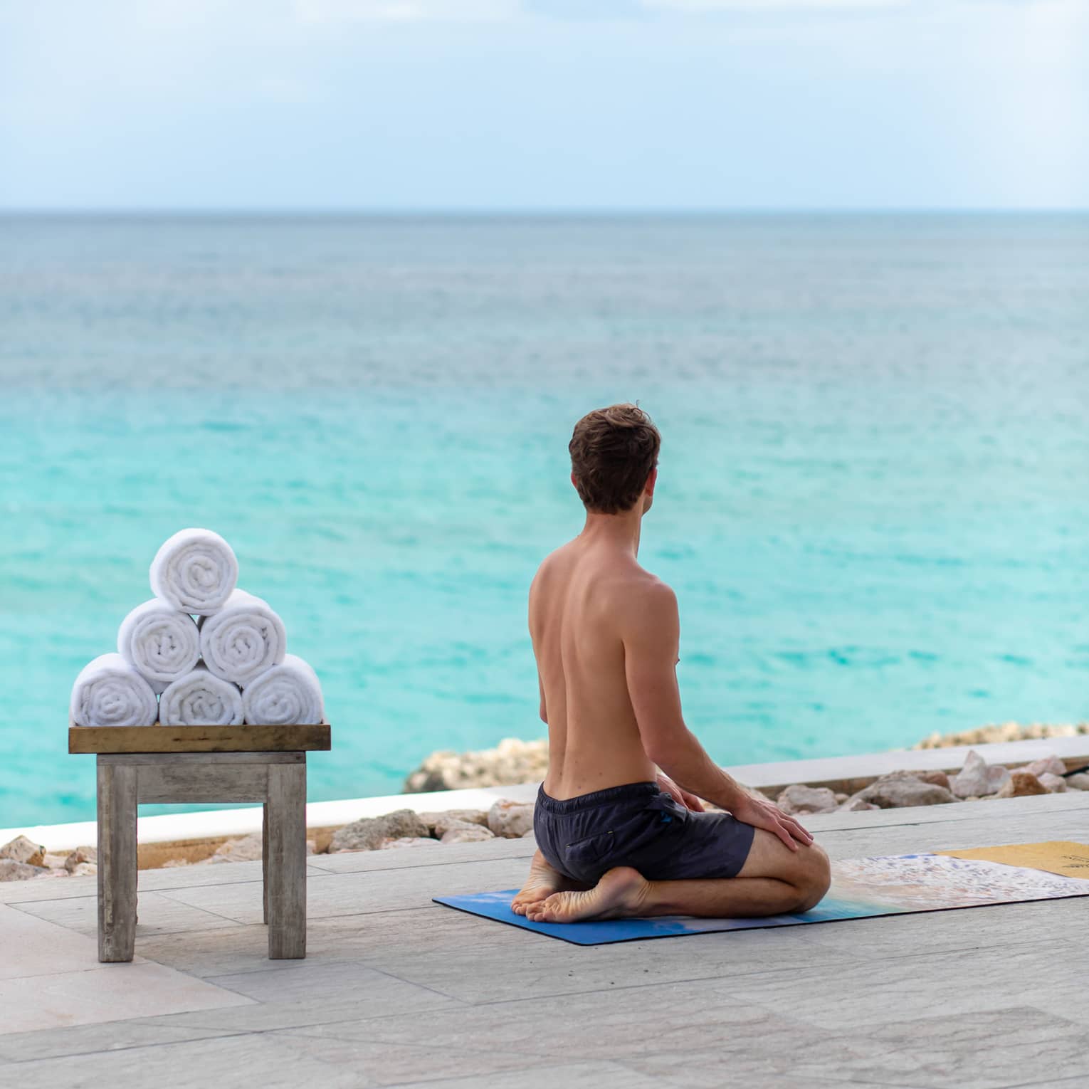 A man sitting on his legs with the ocean in front of him and towels on a bench behind him.