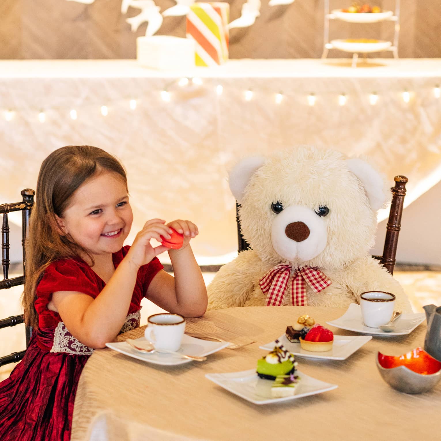 A small girl in a red dress sitting with a large teddy bear at a table with tea and small desserts.