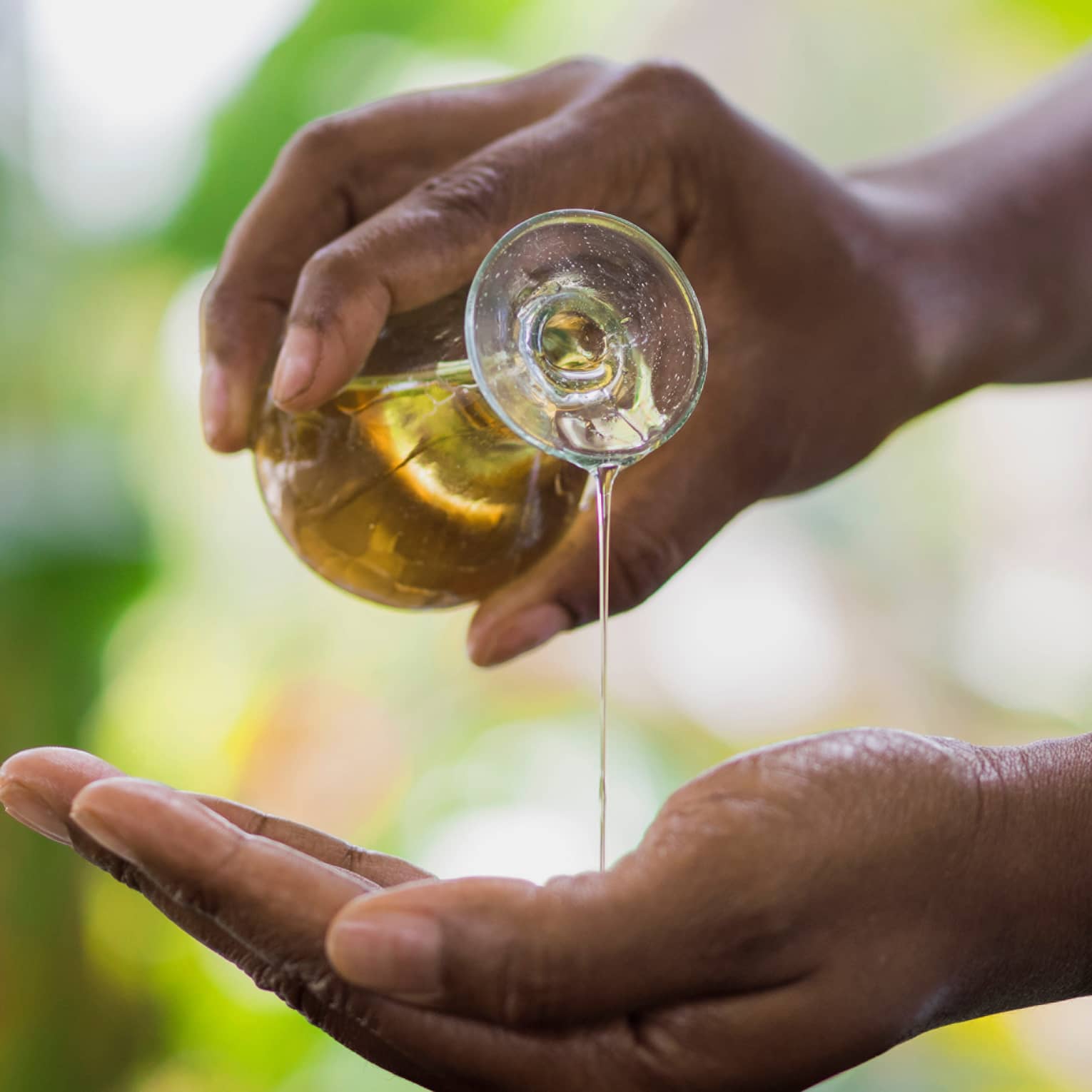 Golden oil flows from a small carafe held in one hand onto the other, a small wooden bowl underneath to catch the overflow.