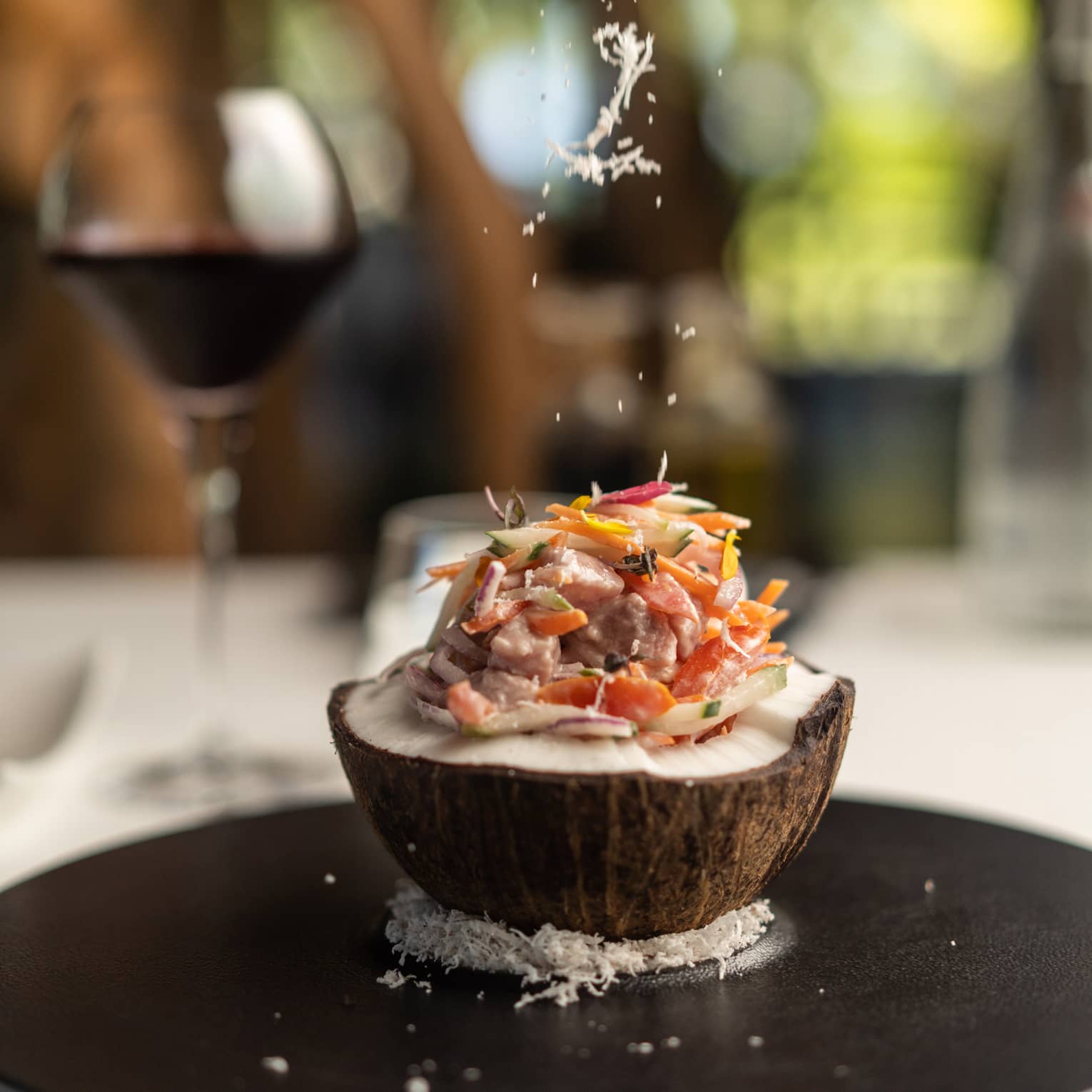 Close-up of an open coconut filled with fish and thinly sliced vegetables, atop a bed of coconut flakes on a round platter.
