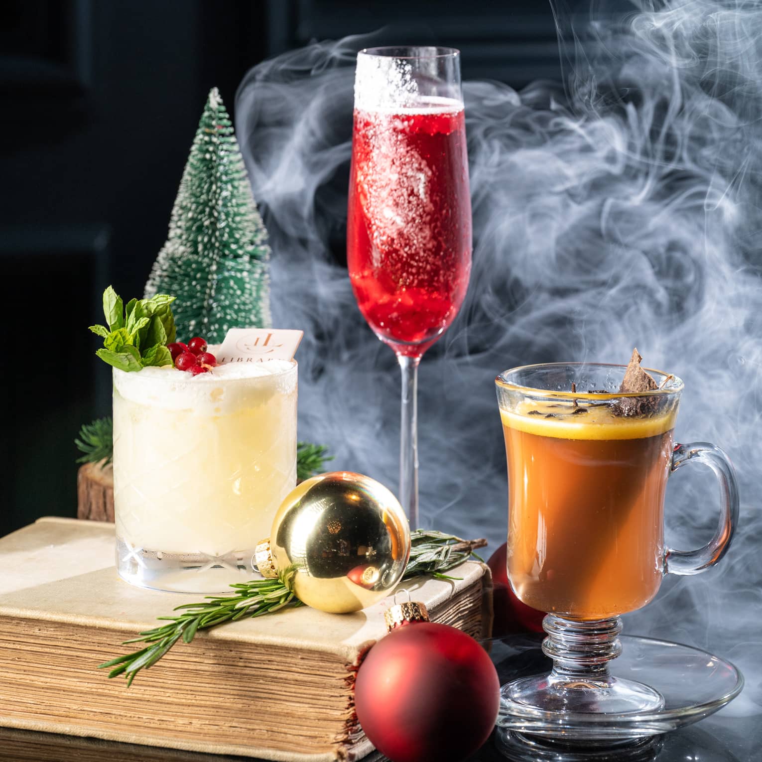 Three festive cocktails arranged on a table with gold and red ornaments, small trees, an old book and a puff of smoke in the background