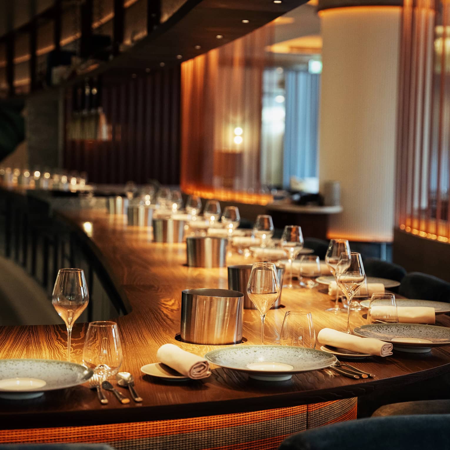 A long, curved solid wood bar with a dozen place settings in warm, golden lighting.