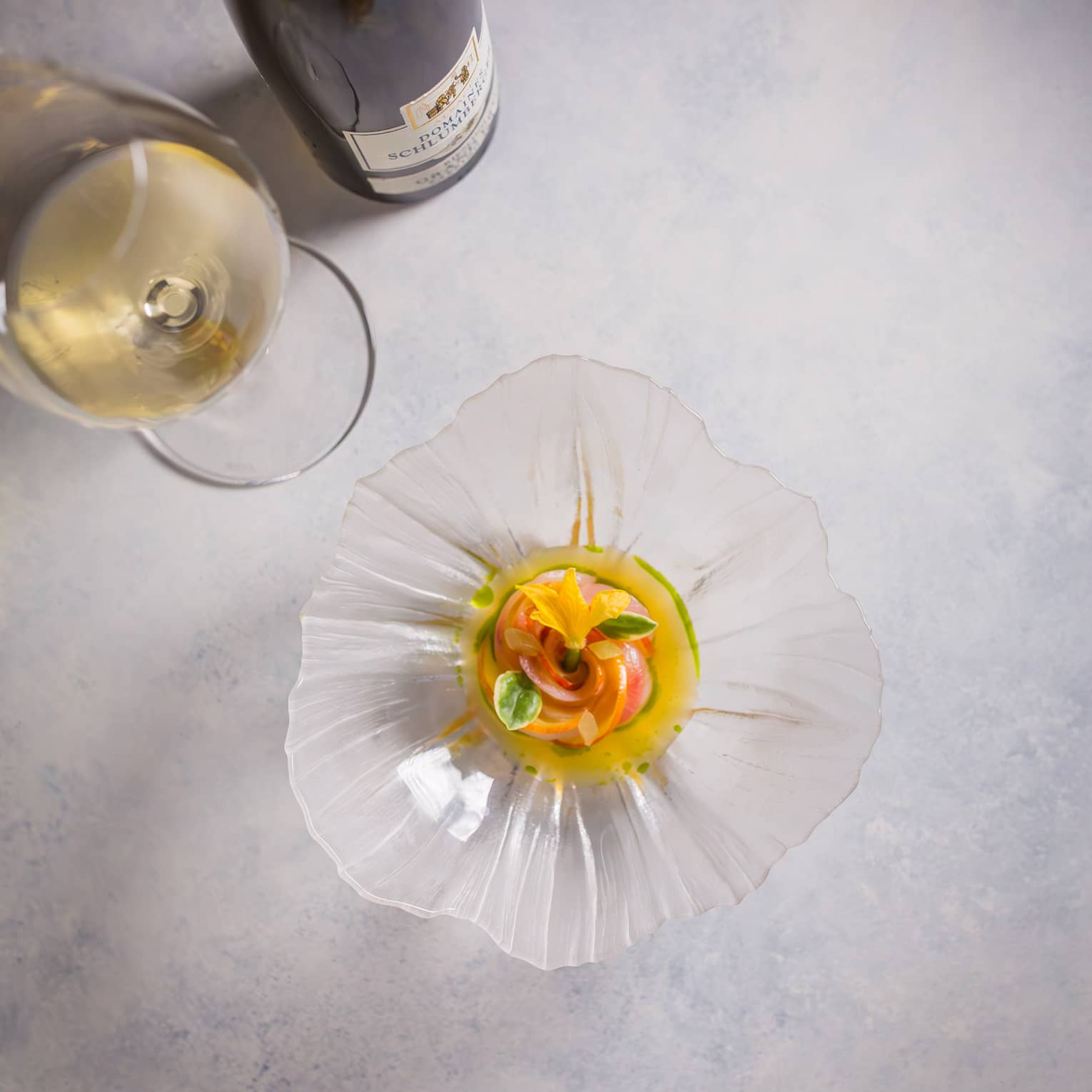 Overhead view of a colourful appetizer garnished with florals in a glass, flower-shaped dish beside a glass of white wine.