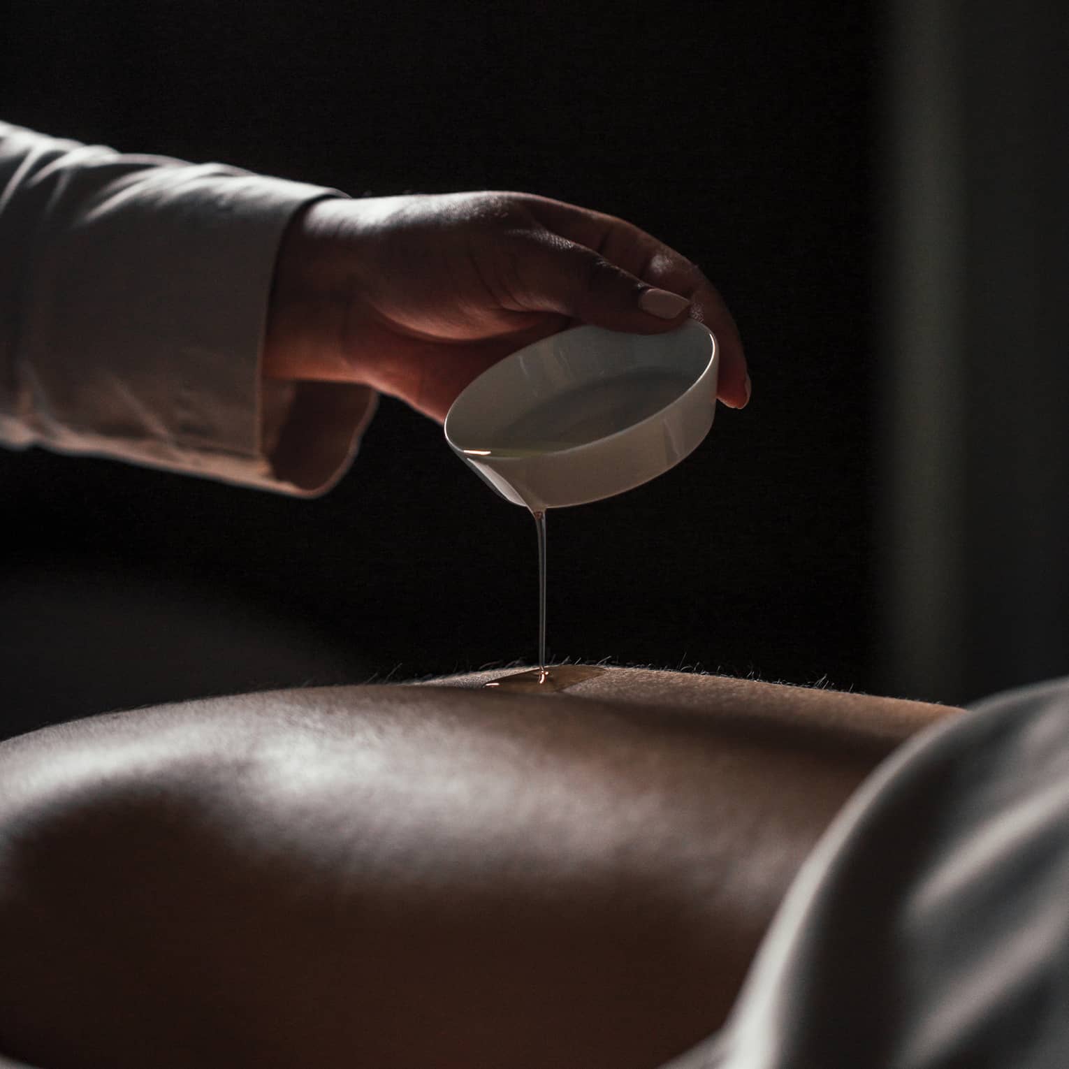 Spa staff pours oil over person's bare back in dark room