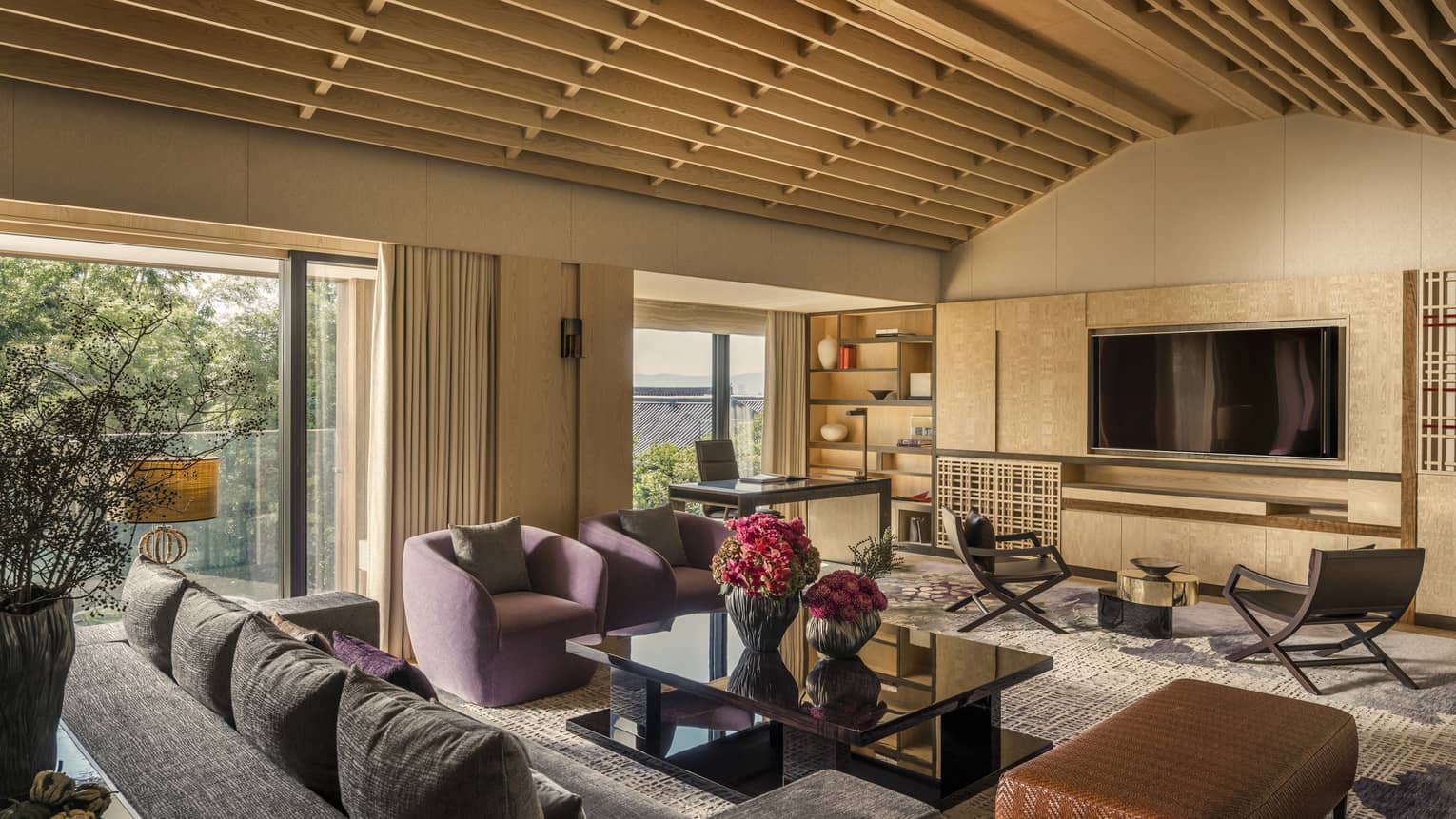 Living area with vaulted ceiling, purple armchairs, sofa, black coffee table, wall-mounted TV, desk, chairs and large windows overlooking greenery
