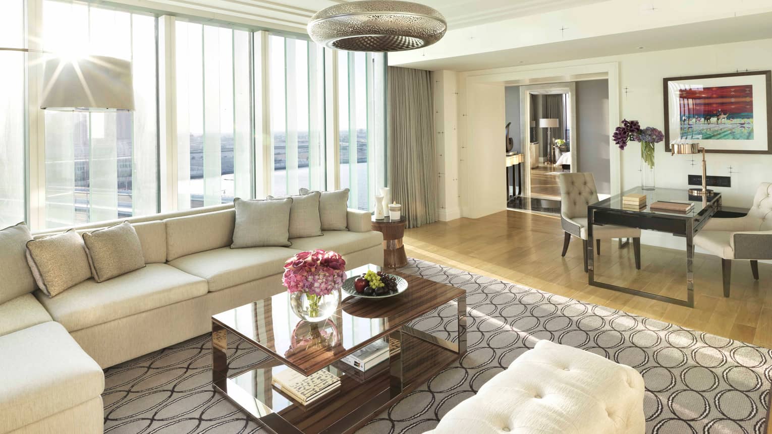 Living room with large grey sectional, rug with octagonal pattern, coffee table and a small dining table to the side, all set infront of a wall of windows