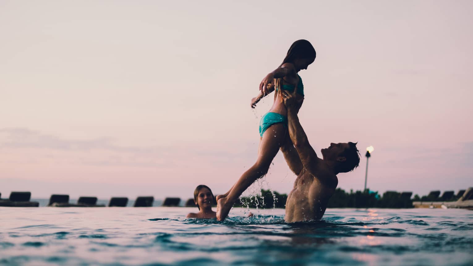 Dad lifts daughter in outdoor swimming pool as smiling boy watches, sunset