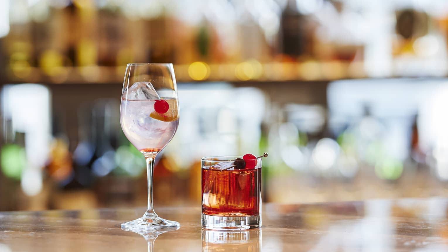 Two cocktails, one is stemmed glass, one in tumbler, with Maraschino cherry garnish on bartop