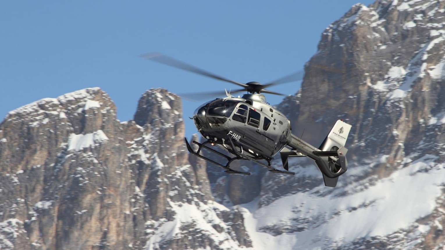 Silver helicopter ascending beside snowy, rocky mountain peaks