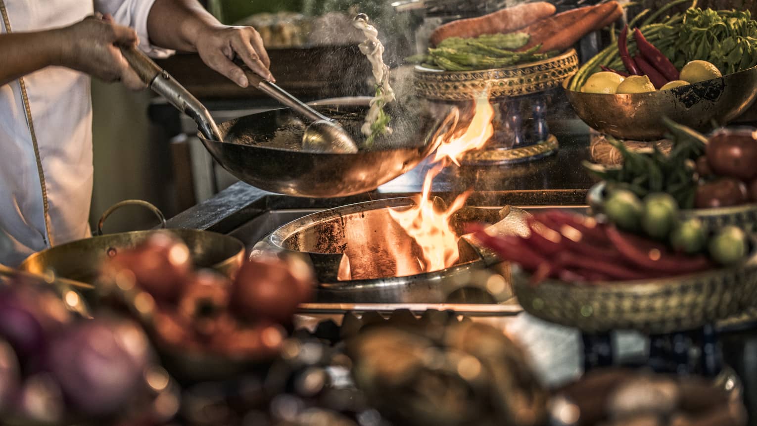 Close-up of chef stirring meals in copper pans over stove, flames 