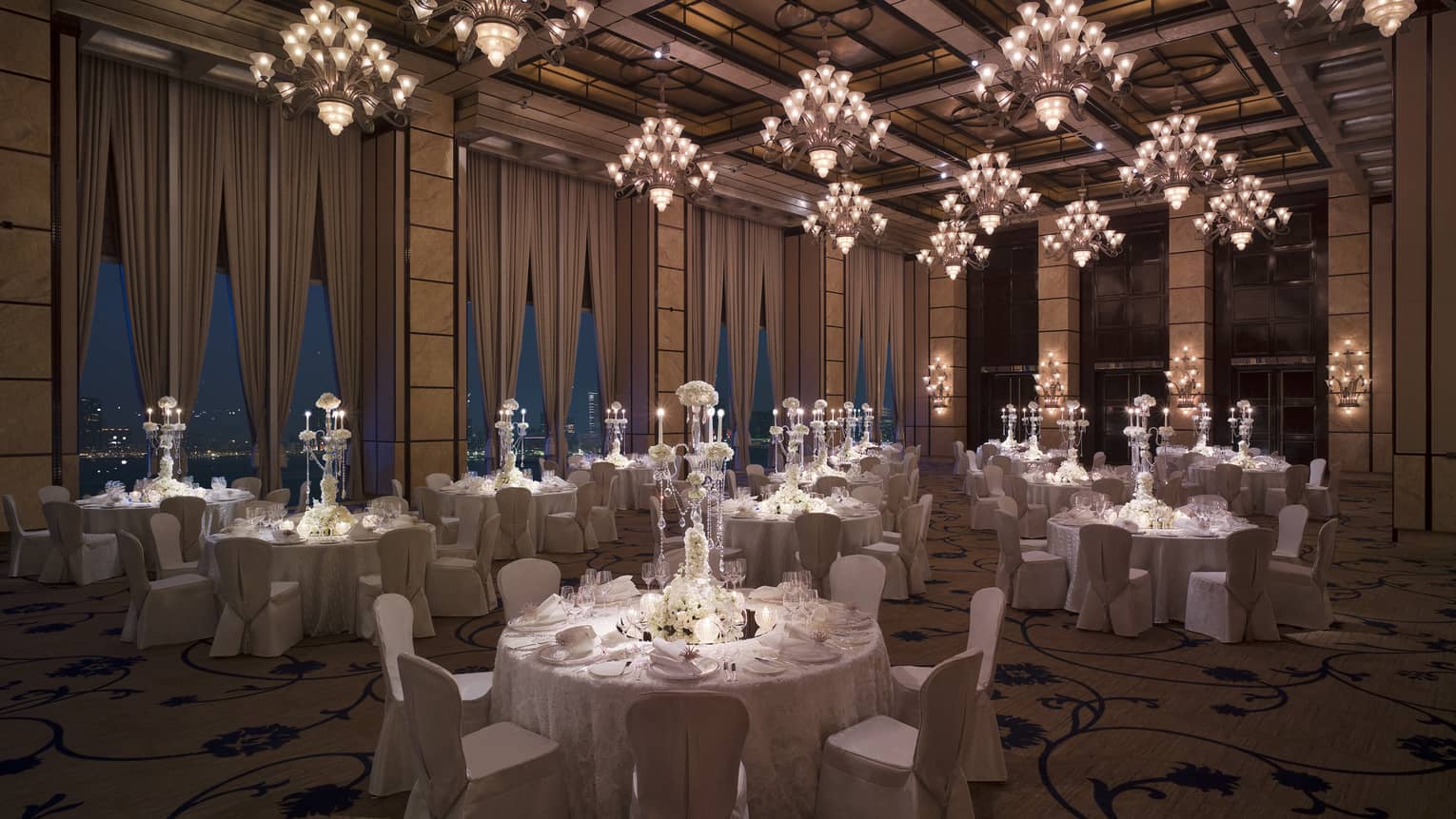 Dimly-lit ballroom with harbor view, round tables with white tablecloths, crystal candelabras, white chairs and floor-to-ceiling windows draped with curtains.