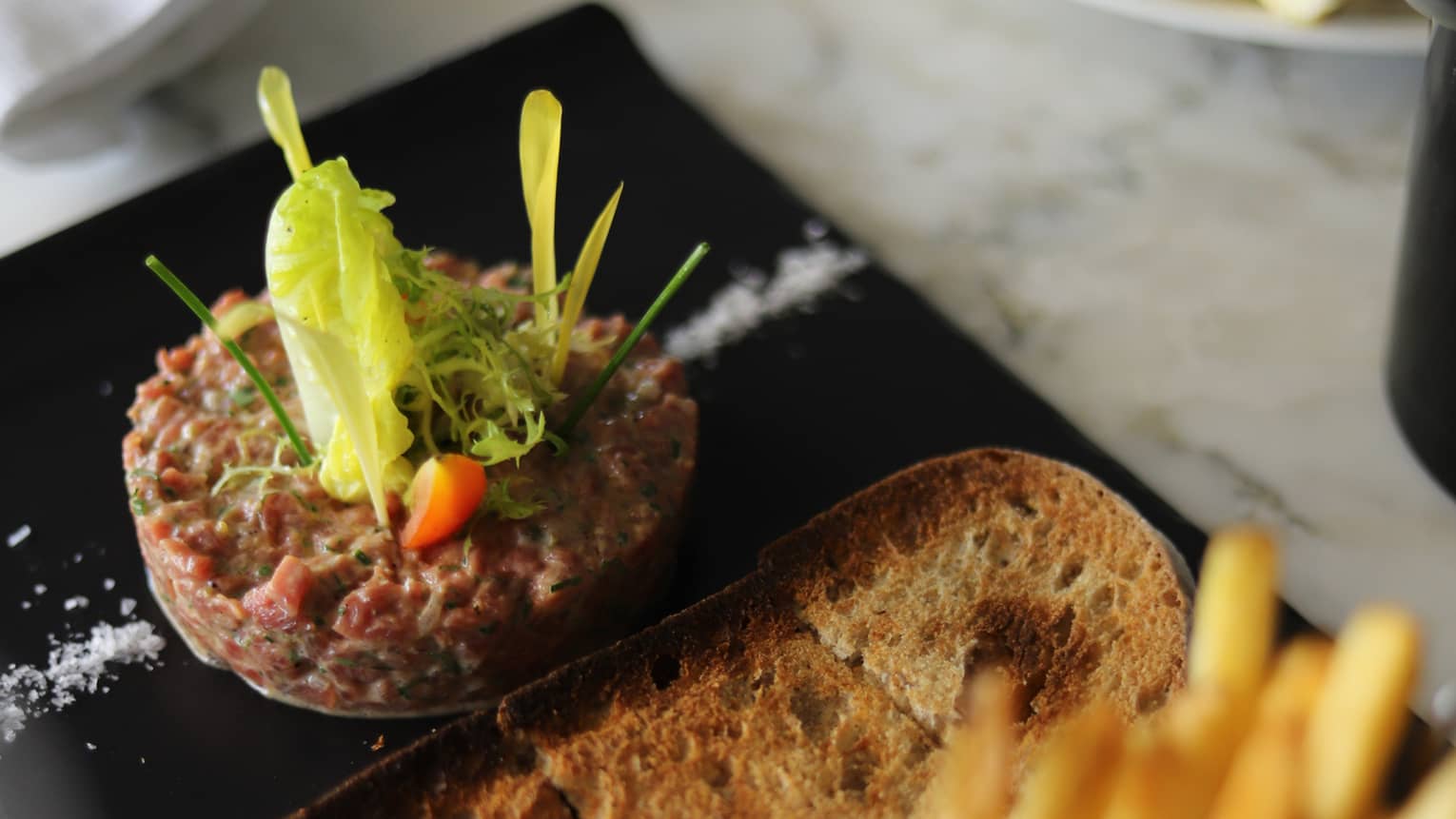 Chopped raw beef tenderloin plated with a slice of toasted sourdough bread, tartare sauce and a small metal basket of French fries