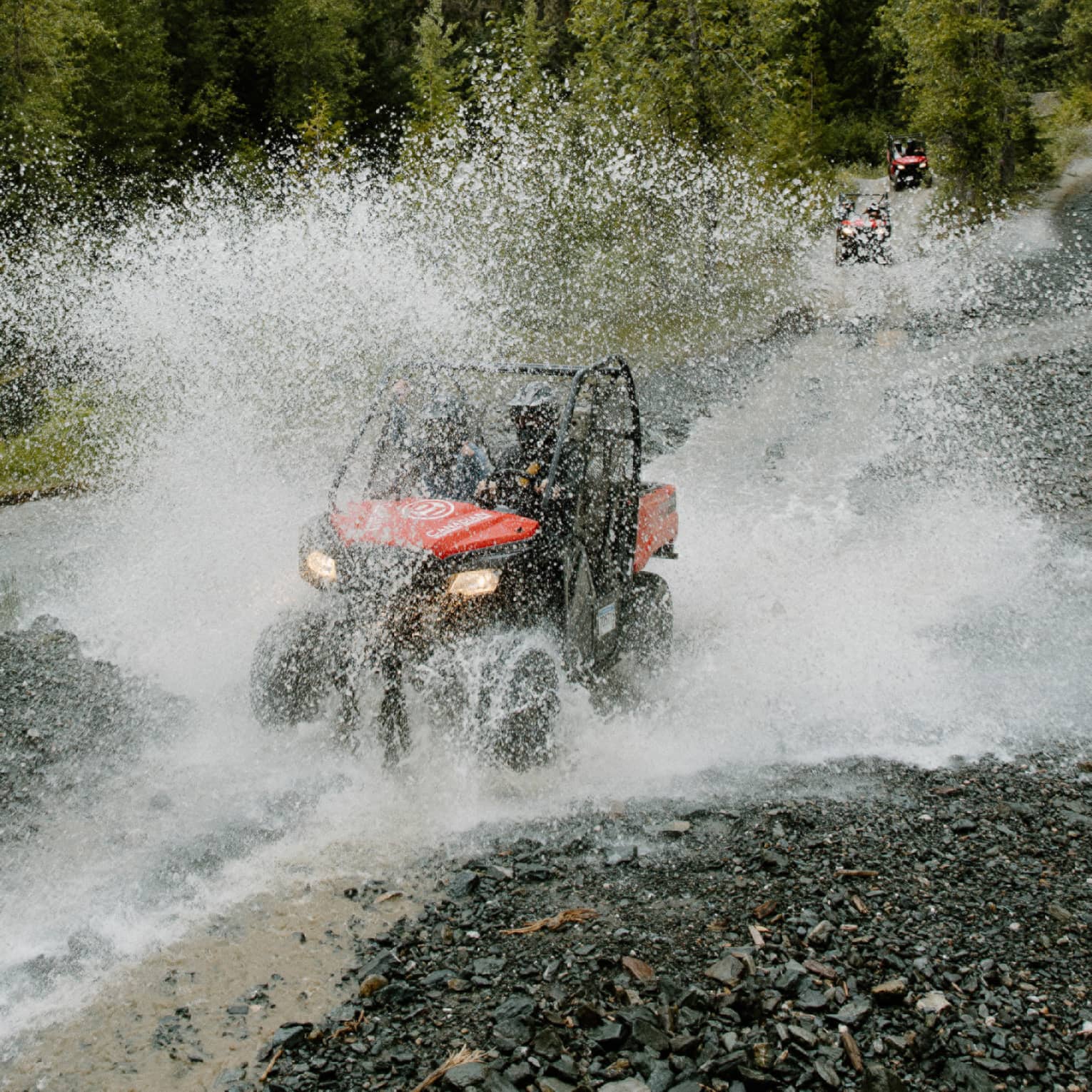 A red-and-black ATV driving through water, creating a thick veil of water spray against a background of pine forest.