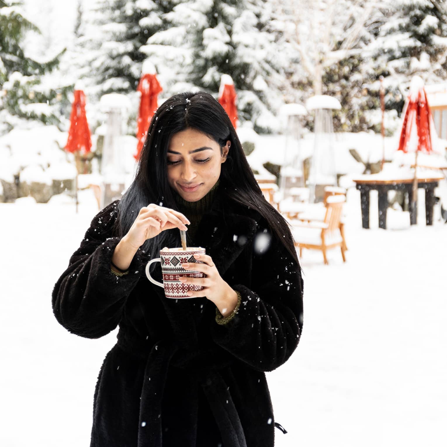 Woman in black coat stirring drink in a festive mug with snow-covered trees and tables in backdrop