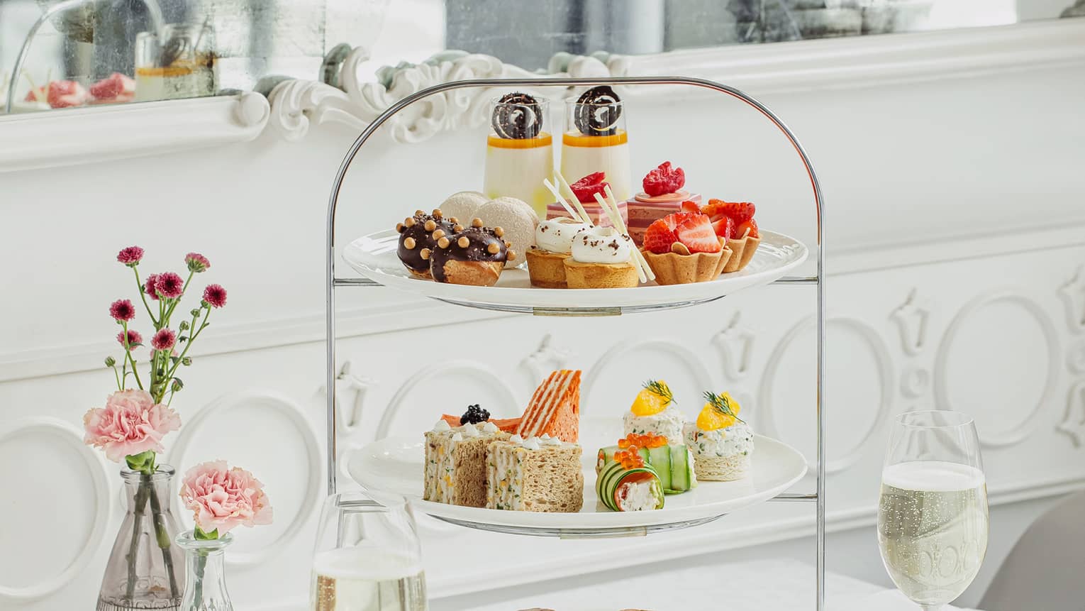 Side dishes, drinks and flowers surround a delicately tiered cake stand holding colourful selections of petite baked goods.