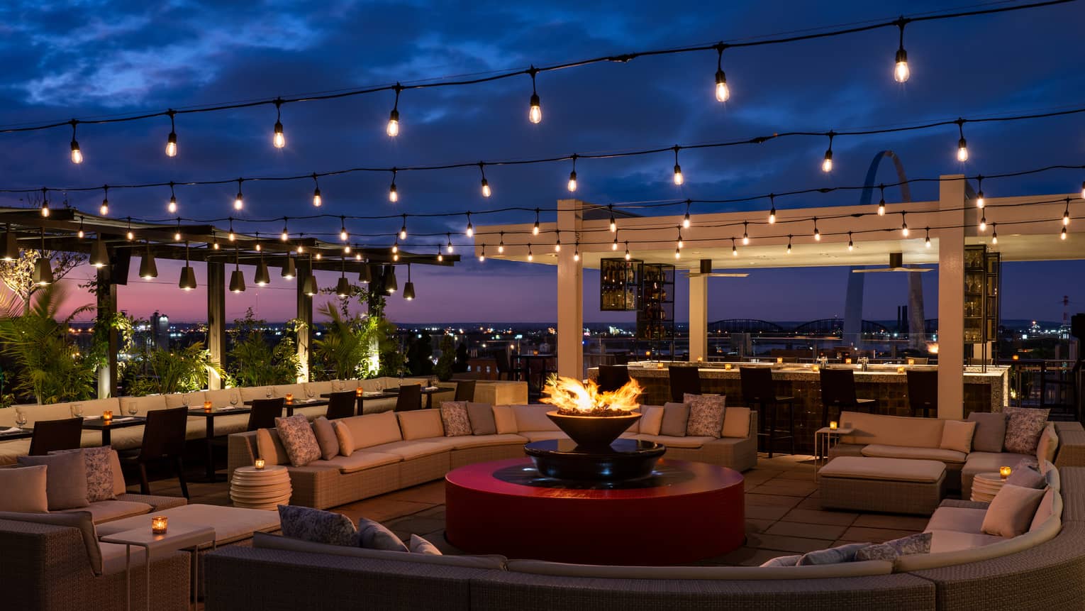 The sky is dark beyond the Cinderhouse Rooftop Bar - decorated with a fire pit, curved sectional, rows of hanging lights and shrubbery