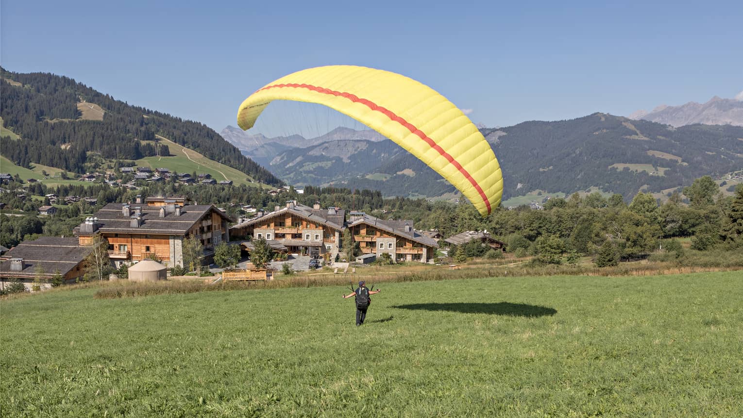 Person on meadow holding yellow parachute in air, facing rustic Megève hotel and verdant mountains