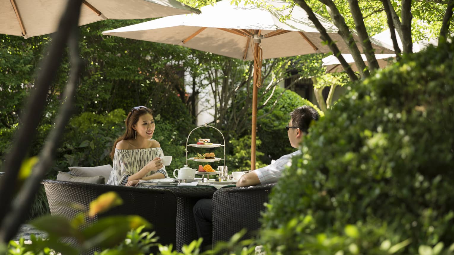 Woman and man drink tea near tray of sweets at table under shade of patio umbrella