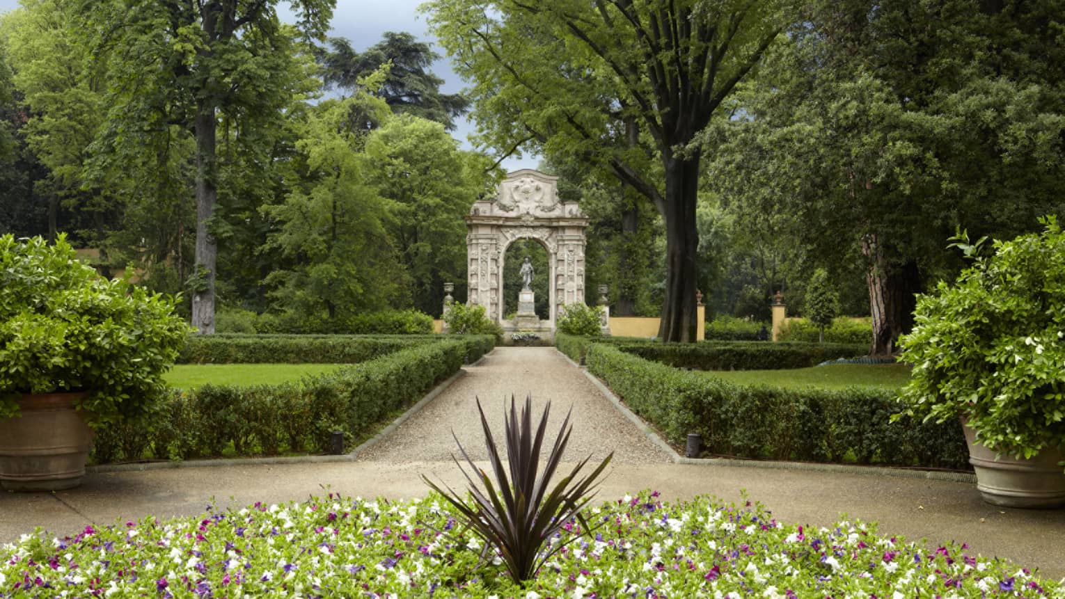 Long garden path lined with shrubs, leading to small arch and stone statue