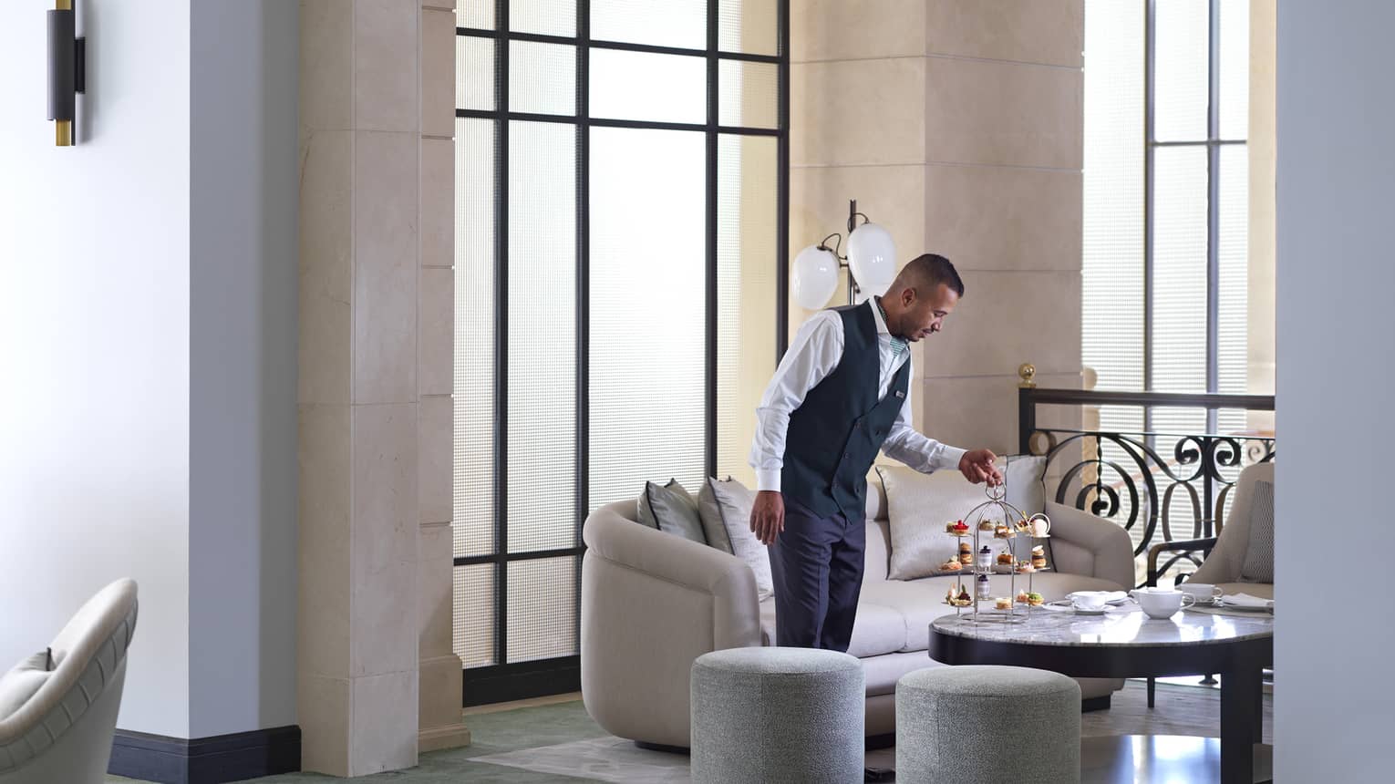 A service member in a navy waistcoat and slacks sets down a three-tiered tray of treats for Afternoon Tea in the Foyer Lounge. 