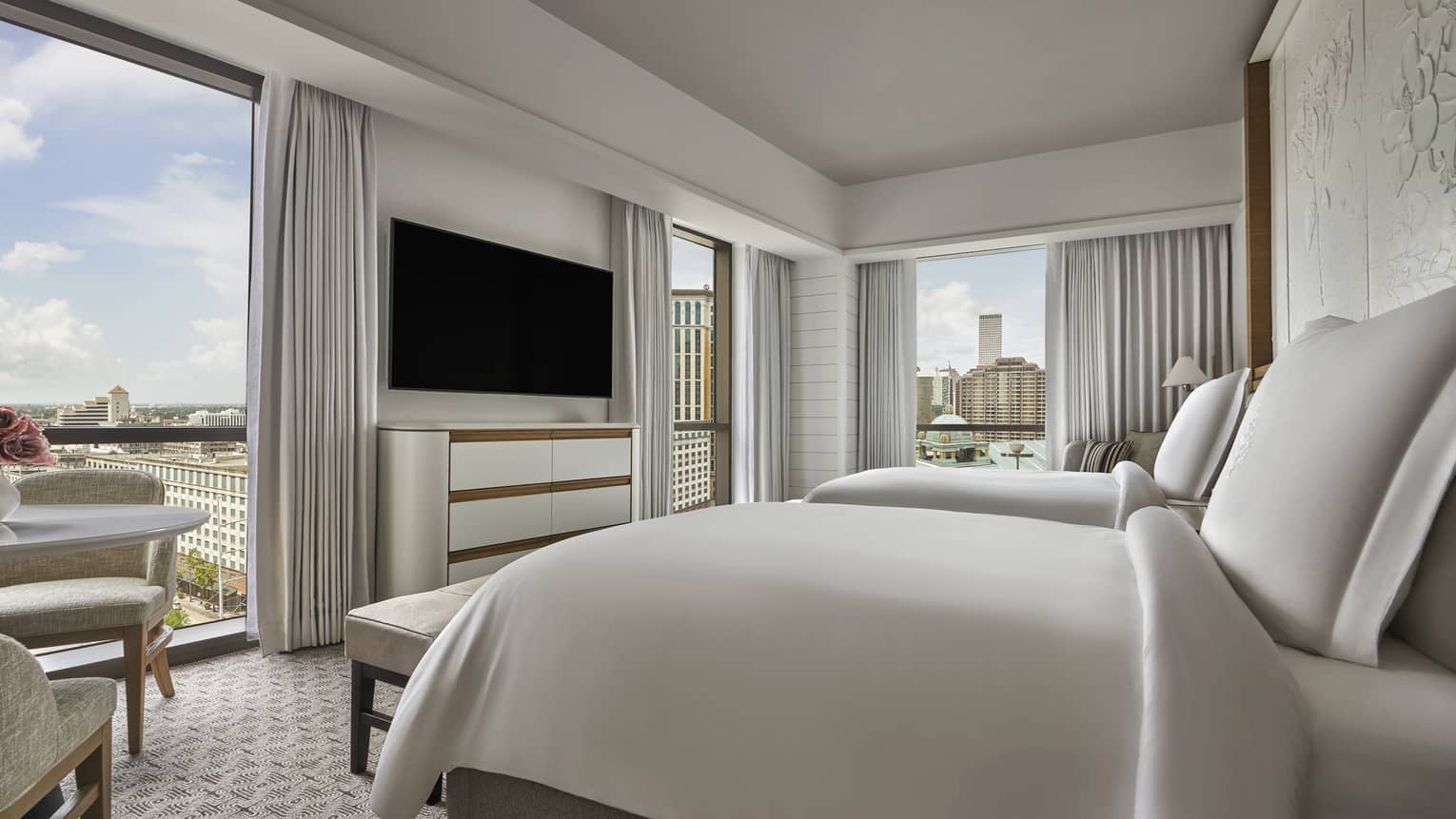 Queen bed with white linens, white curtains and chest of drawers, wall-mounted television, floor-to-ceiling windows with view of downtown New Orleans