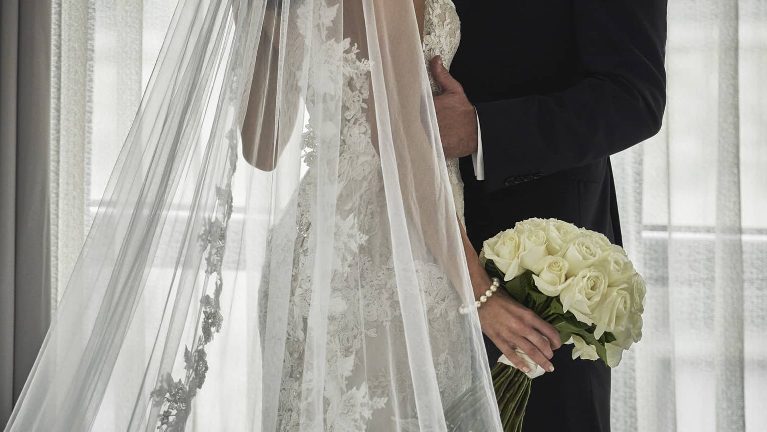 Groom and bride with long lace veil and white wedding bouquet embrace