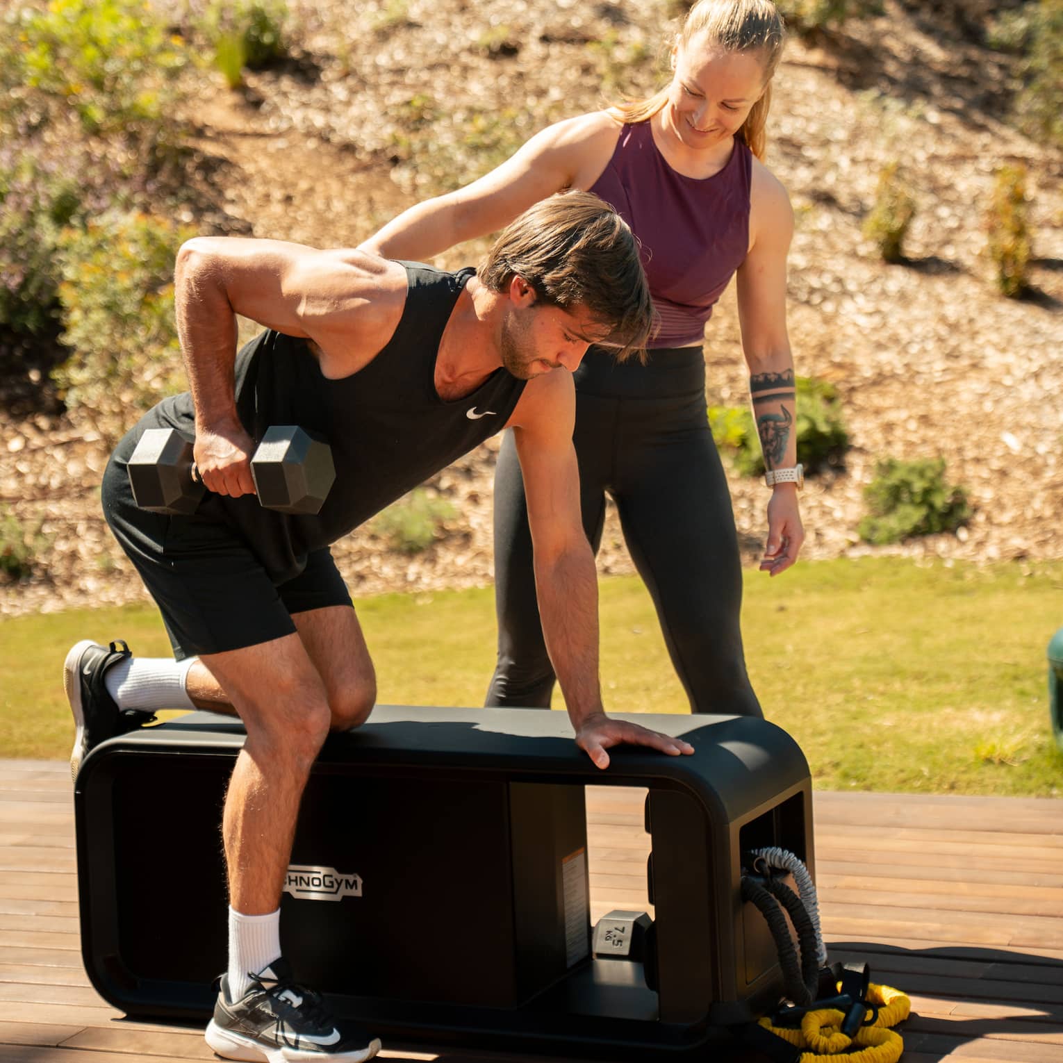 Two people exercise with weights at a workout bench on a wood deck in an outdoor setting.