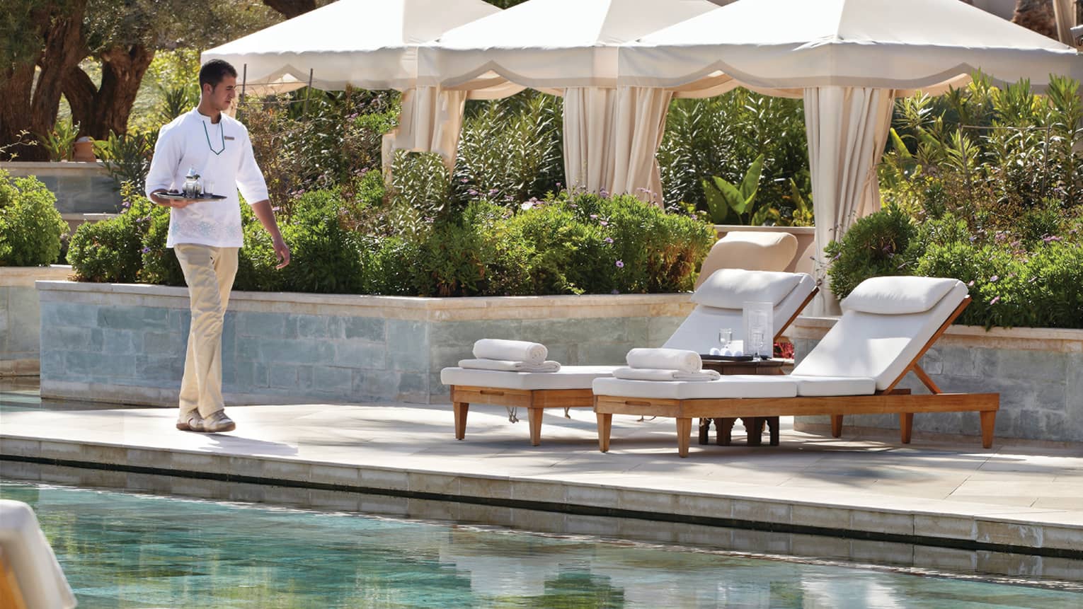 Server carrying tray walks towards two white lounge chairs on swimming pool deck past cabana