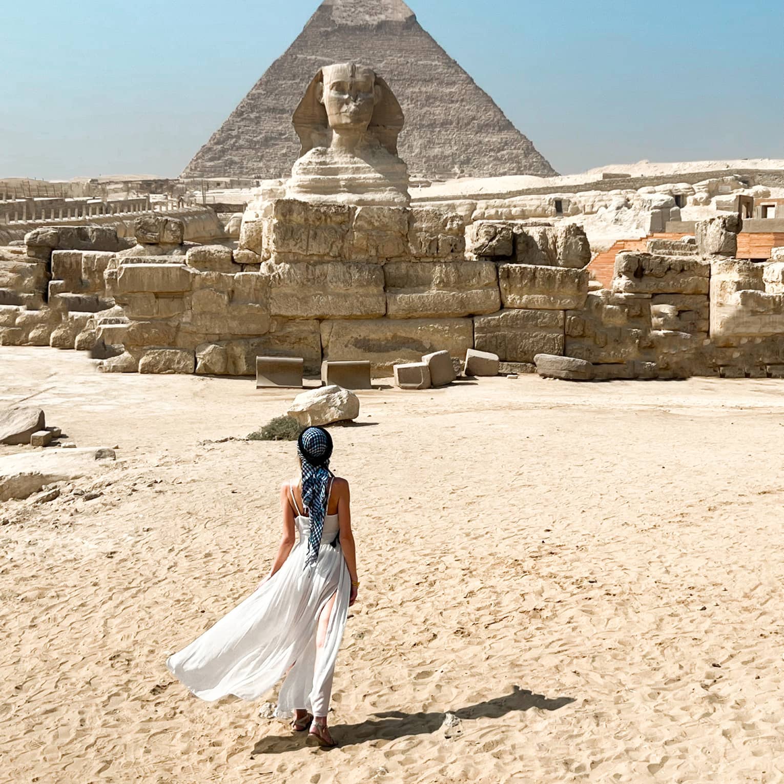 View from behind as a guest crosses golden desert sand toward the immense ruins and sphinx, a pyramid rising up beyond.