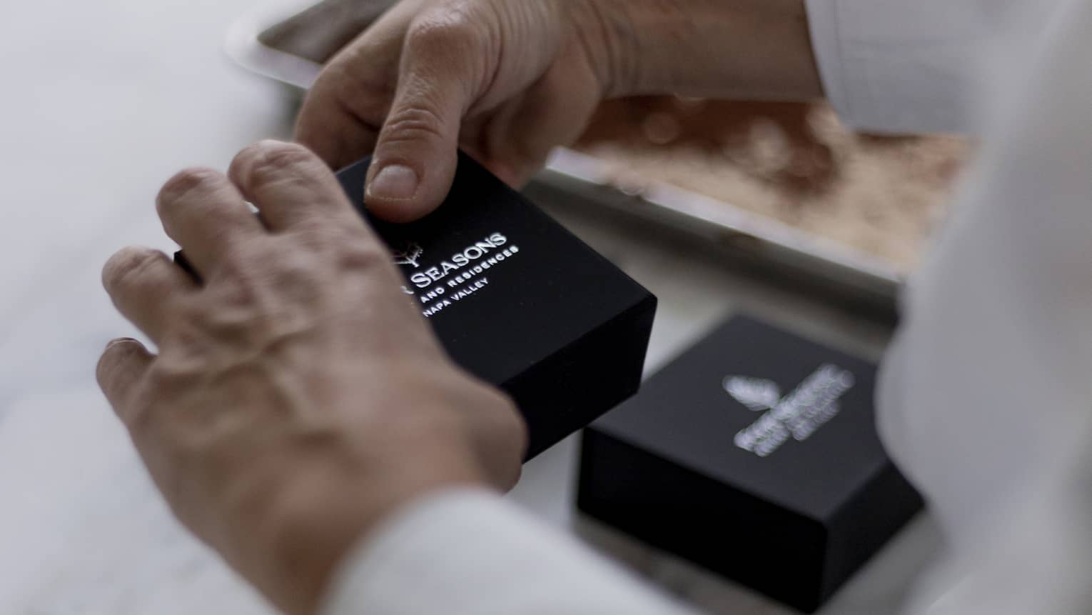 Hands holding a small, black box with the Four Seasons name and logo in silver; a blurred cookie sheet in the background.