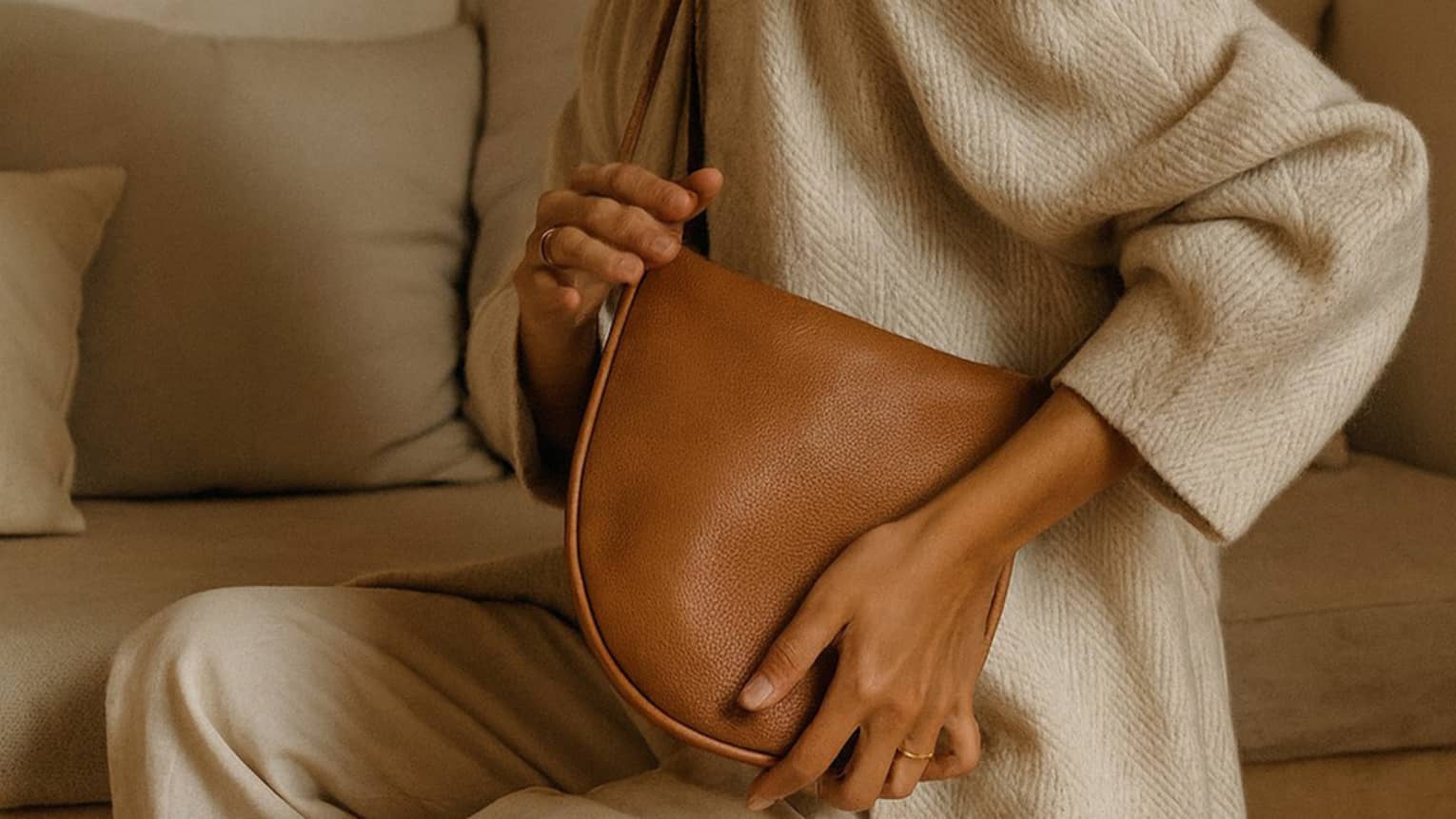A woman dressed in a beige linen jacket and pants poses with a brown leather messenger bag